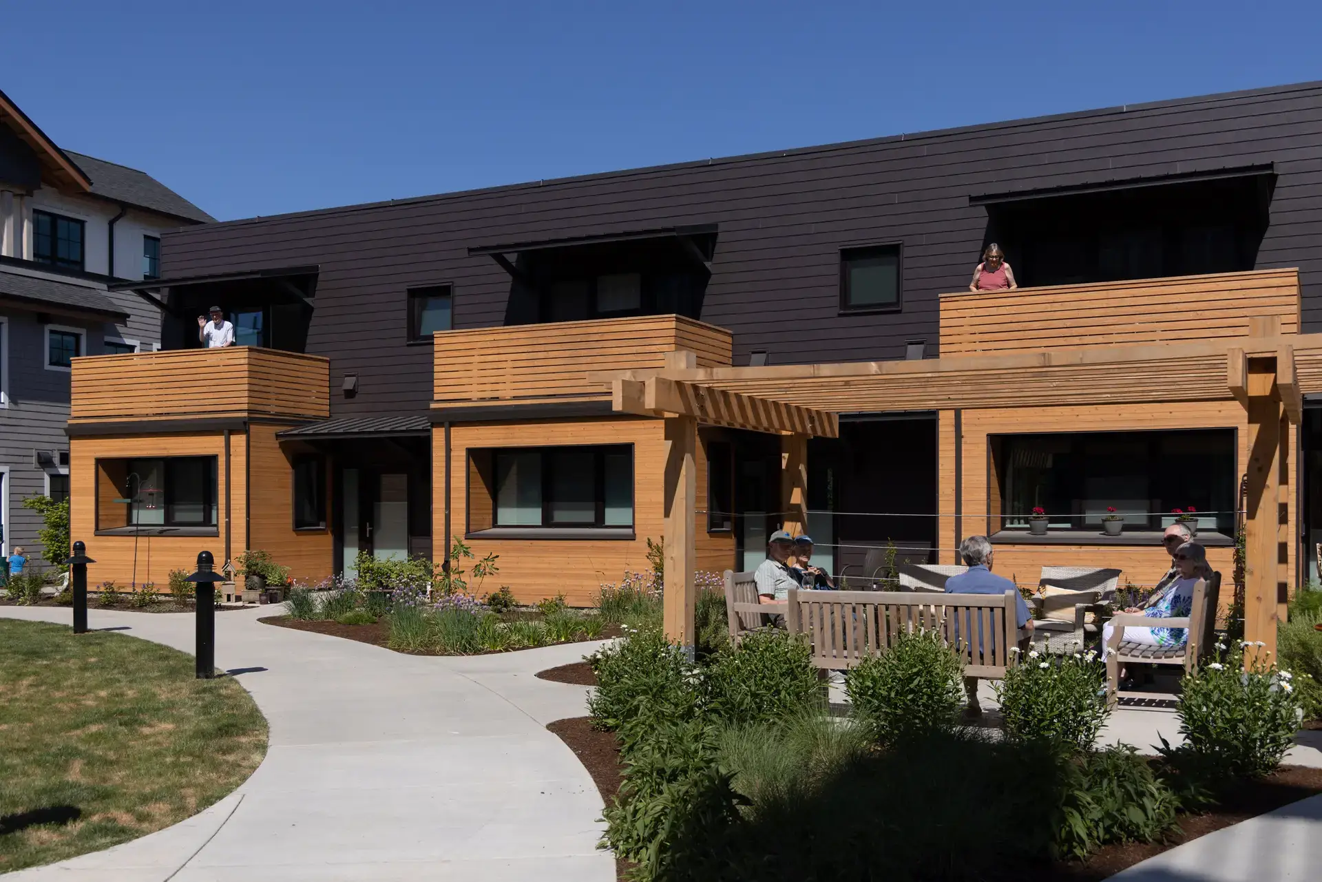 Courtyard view of Trillium Townhomes, oriented around a central pedestrian path and a landscaped courtyard. A wooden pergola provides a shaded seating area where several residents are seen gathering, reflecting the community's focus on social connection.