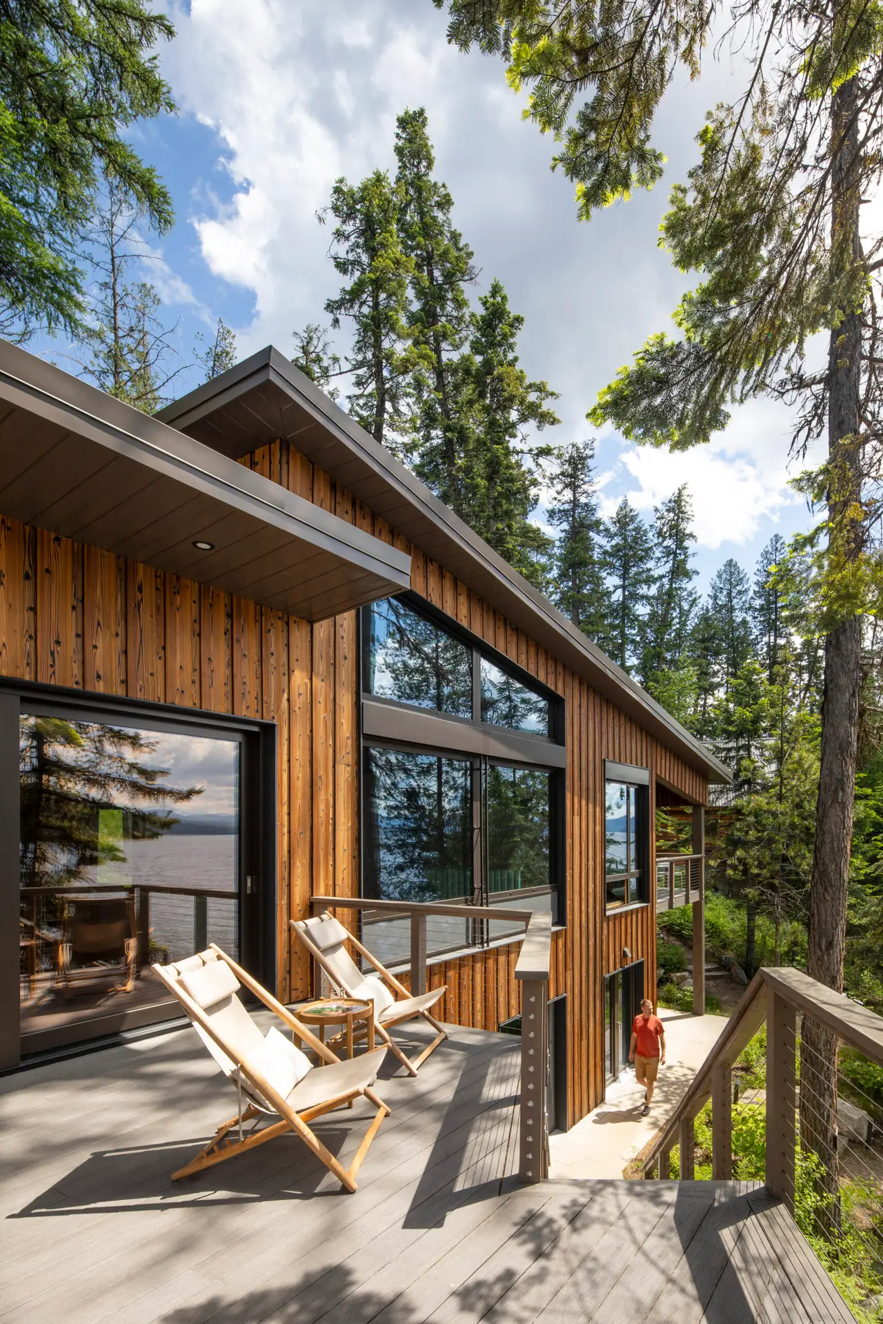 Priest Lake Cabin deck with patio chairs in the foreground and a lower portion of deck accessed by stairs. The cabin is in the frame and has wood siding and dark, reflective windows.
