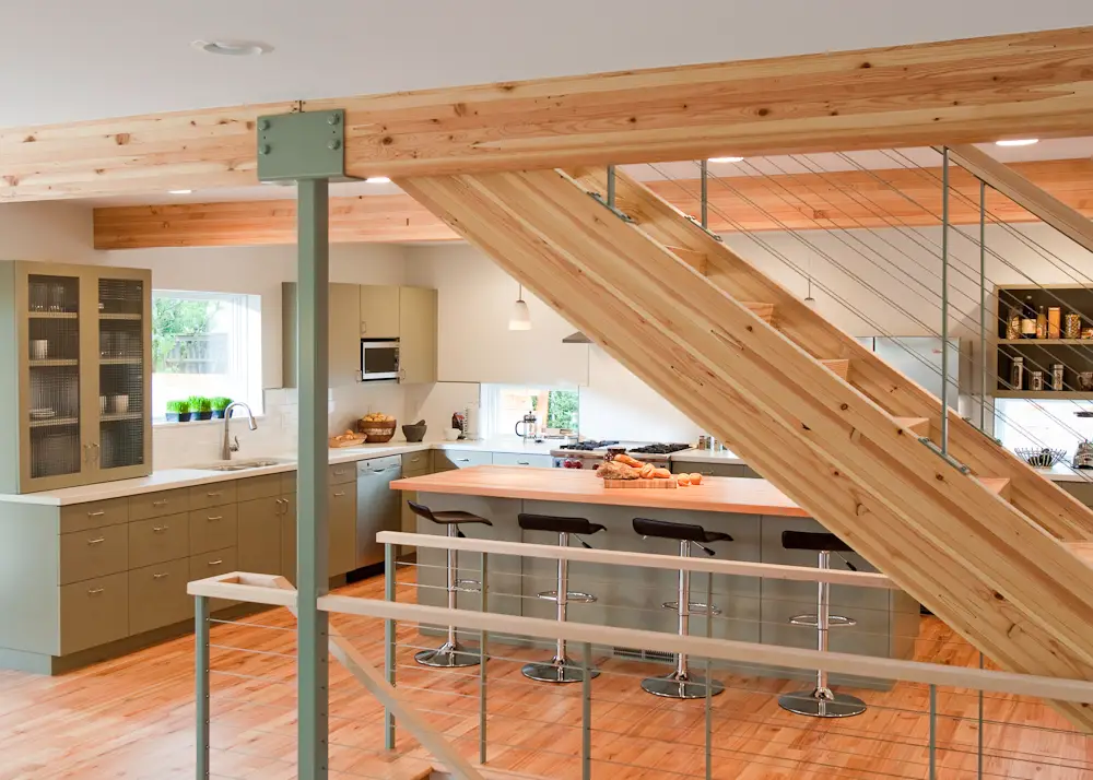 The kitchen and of the Hale Residence featuring a central open-riser staircase with thick wood treads and a modern cable railing system that maintains sightlines and allows natural light to flow through the space. The structure utilizes exposed Glulam beams and a prominent industrial-style steel post to support the open floor plan. Flooring: The home is anchored by wide-plank natural wood flooring that coordinates with the ceiling beams and stair treads