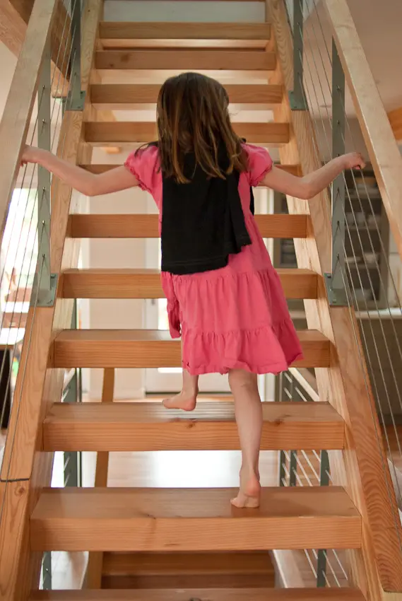 A child climbing a modern, open-riser staircase. The stairs are built with thick, solid wood treads and open risers, which allow light to pass through and maintain an airy, spacious feel within the home's high-performance layout. The staircase is flanked by a contemporary cable railing system. This features slim, vertical steel posts and horizontal stainless steel cables, providing safety while minimizing visual obstruction. Consistent with Green Hammer's commitment to sustainability, the wood used for the treads and stringers is likely FSC-certified