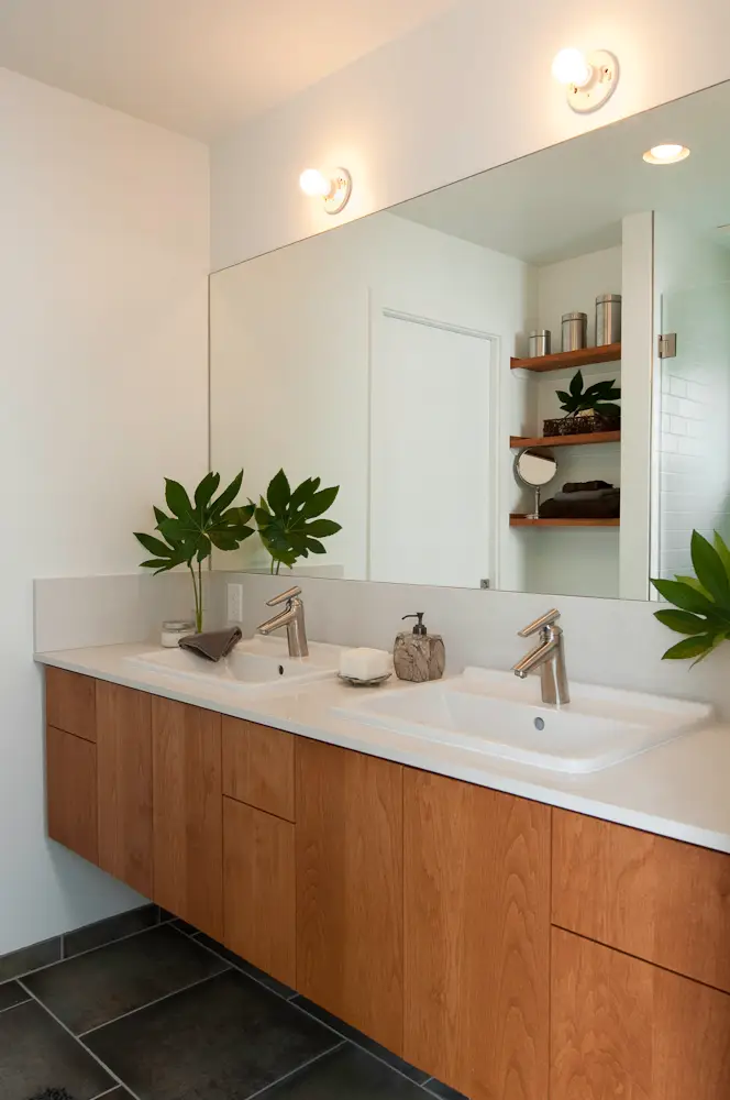 A contemporary primary bathroom of the Hale Residence featuring a long, wall-mounted vanity crafted from light-toned wood with a prominent vertical grain. The vanity is topped with a sleek white quartz countertop and a matching low-profile backsplash. It features two large rectangular undermount sinks with contemporary brushed-nickel or chrome faucets. The floor is clad in large-format slate-grey tiles with subtle natural textures, providing a cool-toned contrast to the warm wood of the vanity. A massive, unframed rectangular mirror spans the entire width of the vanity, reflecting light to brighten the room. Two minimalist, globe-style vanity lights are mounted directly above the mirror, providing even illumination.