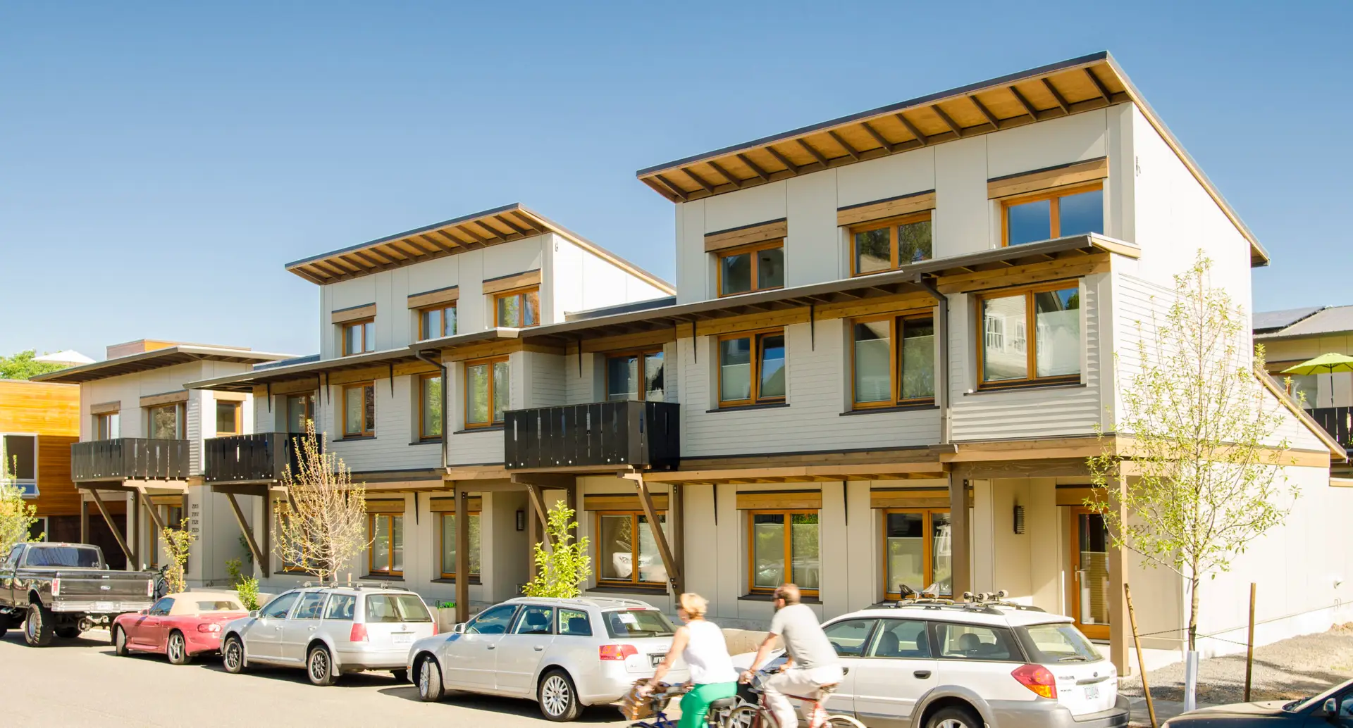 A view of Ankeny Row Zero Energy cohousing from the street showing the exterior shading, balconies and expressed wood finishes. The architecture is a contemporary take on the traditional craftsman style typical of the surrounding neighborhood, featuring light-colored siding and warm wood-framed windows.