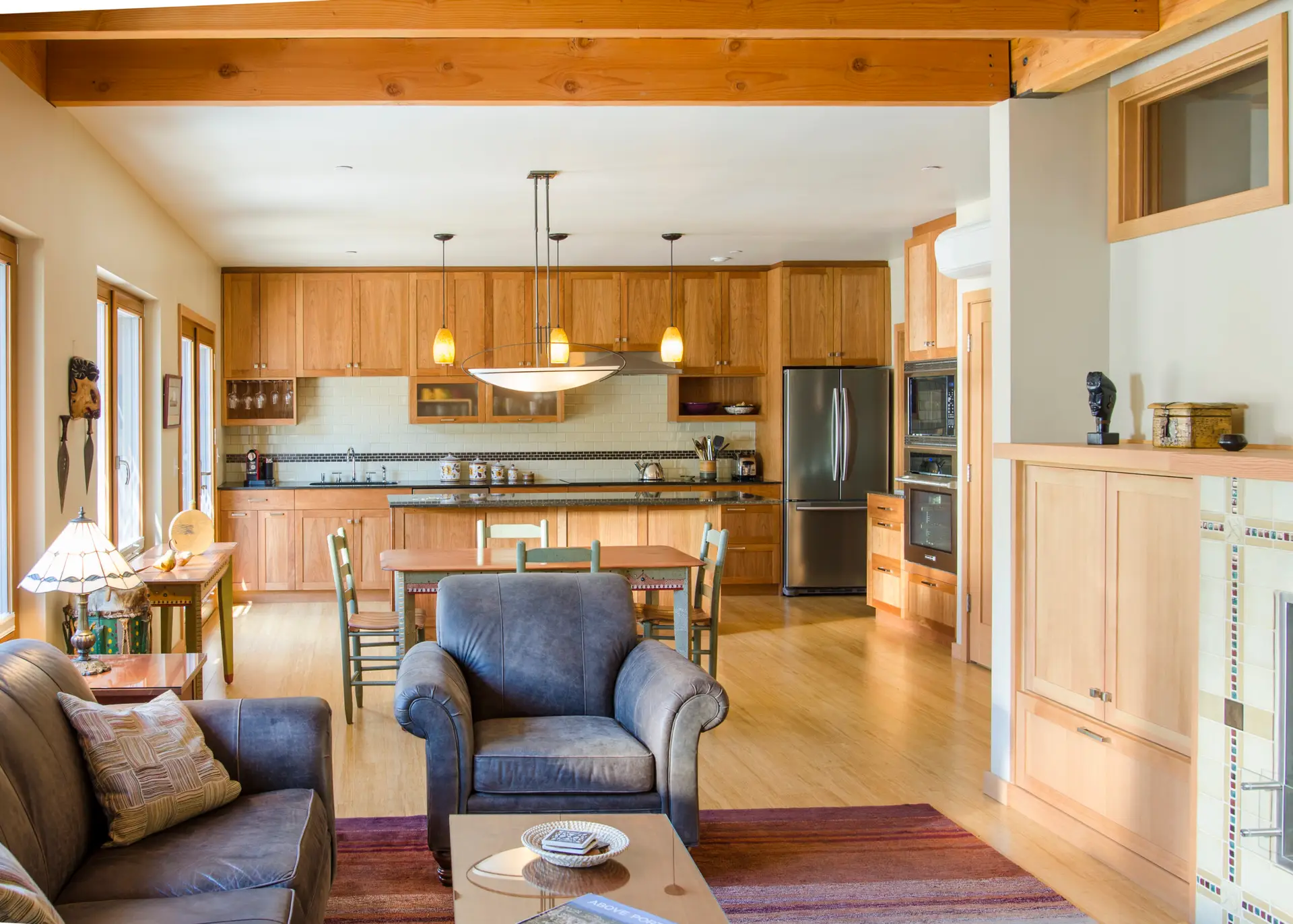 An open-concept living, dining, and kitchen area of a unit in Ankeny Row, a net-zero energy cohousing community in Portland, Oregon. The space is designed with a warm, "modern craftsman" aesthetic that prioritizes natural materials and accessibility. Exposed heavy timber beams span the ceiling, adding a sense of structure and warmth. Large, wood-framed windows and a glass door on the left allow for ample natural light and a view of the outdoors.