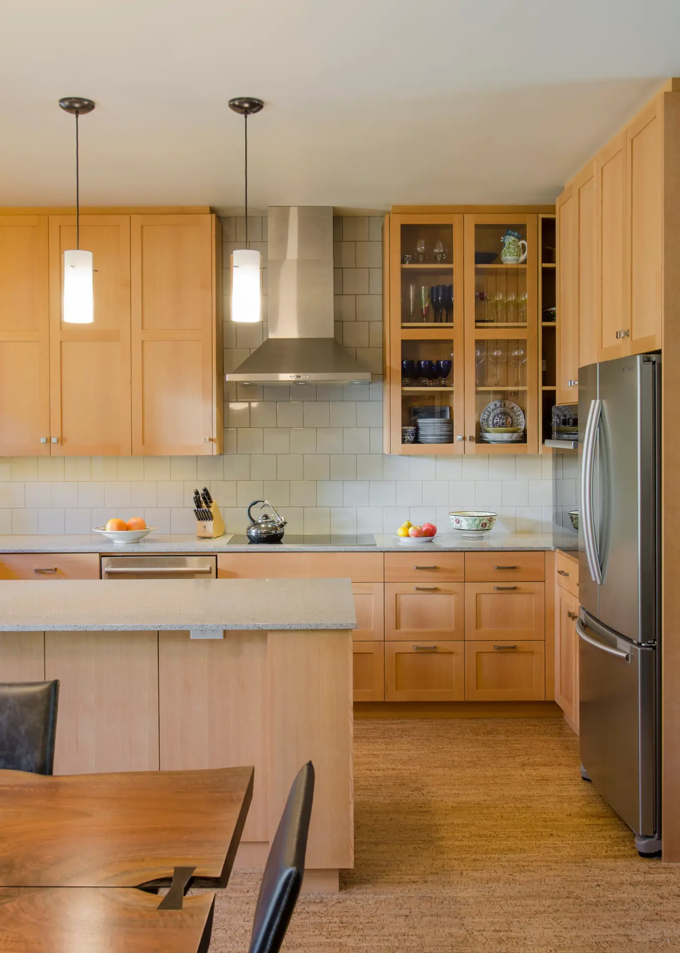A modern, sustainably designed kitchen at Ankeny Row, a net-zero energy cohousing community in Portland, Oregon, designed and built by Green Hammer. The kitchen is anchored by custom light maple cabinets with a clean, horizontal grain. Some upper units feature glass fronts to display glassware. The floor is made of cork, chosen for its sustainability, durability, and comfort underfoot—materials often used in Green Hammer's high-performance projects.