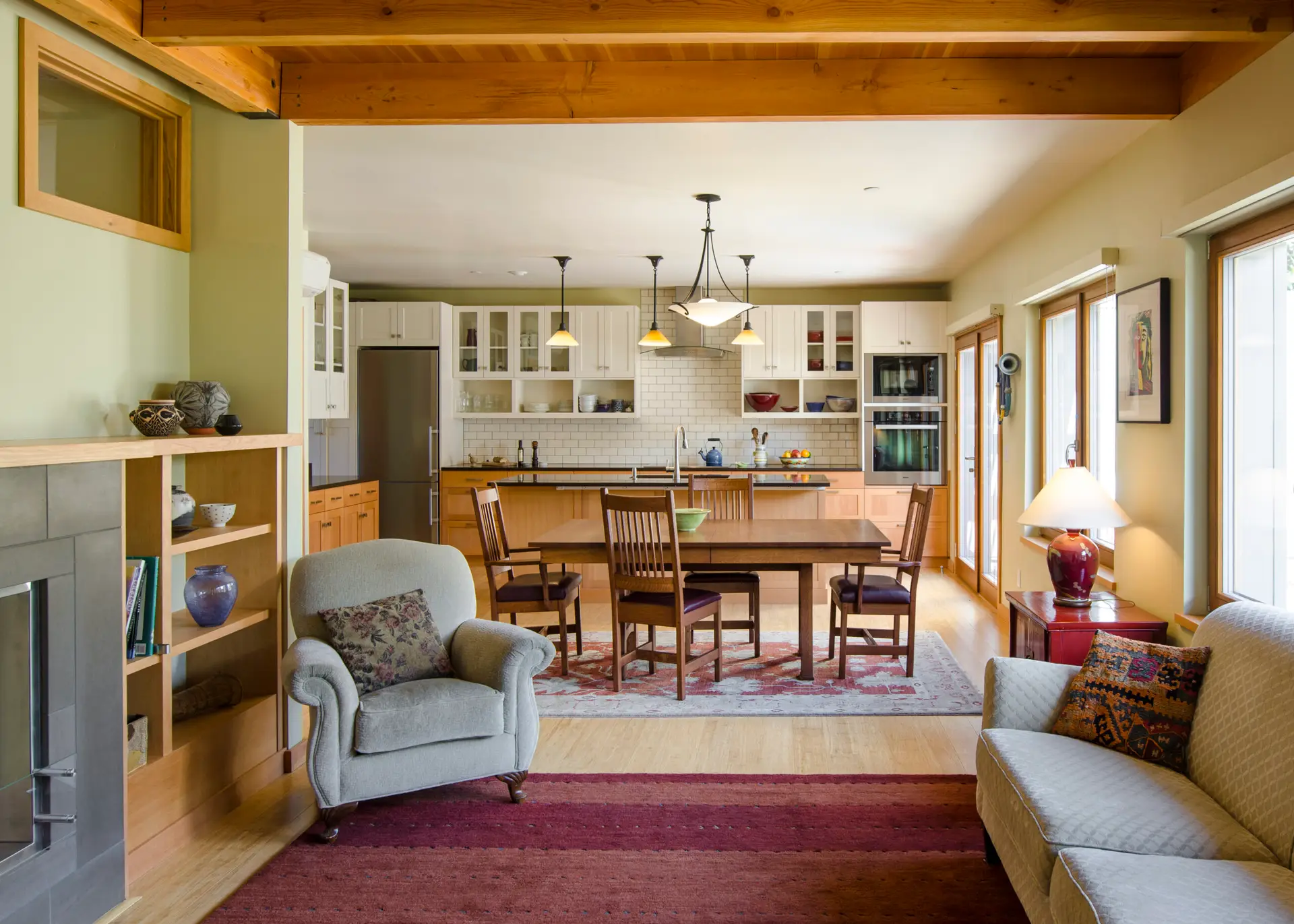 An open-concept living, dining, and kitchen area of a home in Ankeny Row, a net-zero energy cohousing community in Portland, Oregon. The space features a modern craftsman aesthetic, combining clean lines with warm, natural materials. In the foreground, a cozy sitting area is anchored by a grey armchair and a matching sofa. Two layered area rugs—one a solid deep burgundy and the other a patterned traditional style—add warmth to the light wood flooring. A fireplace with a grey tile surround and a light wood mantel is situated on the left.
