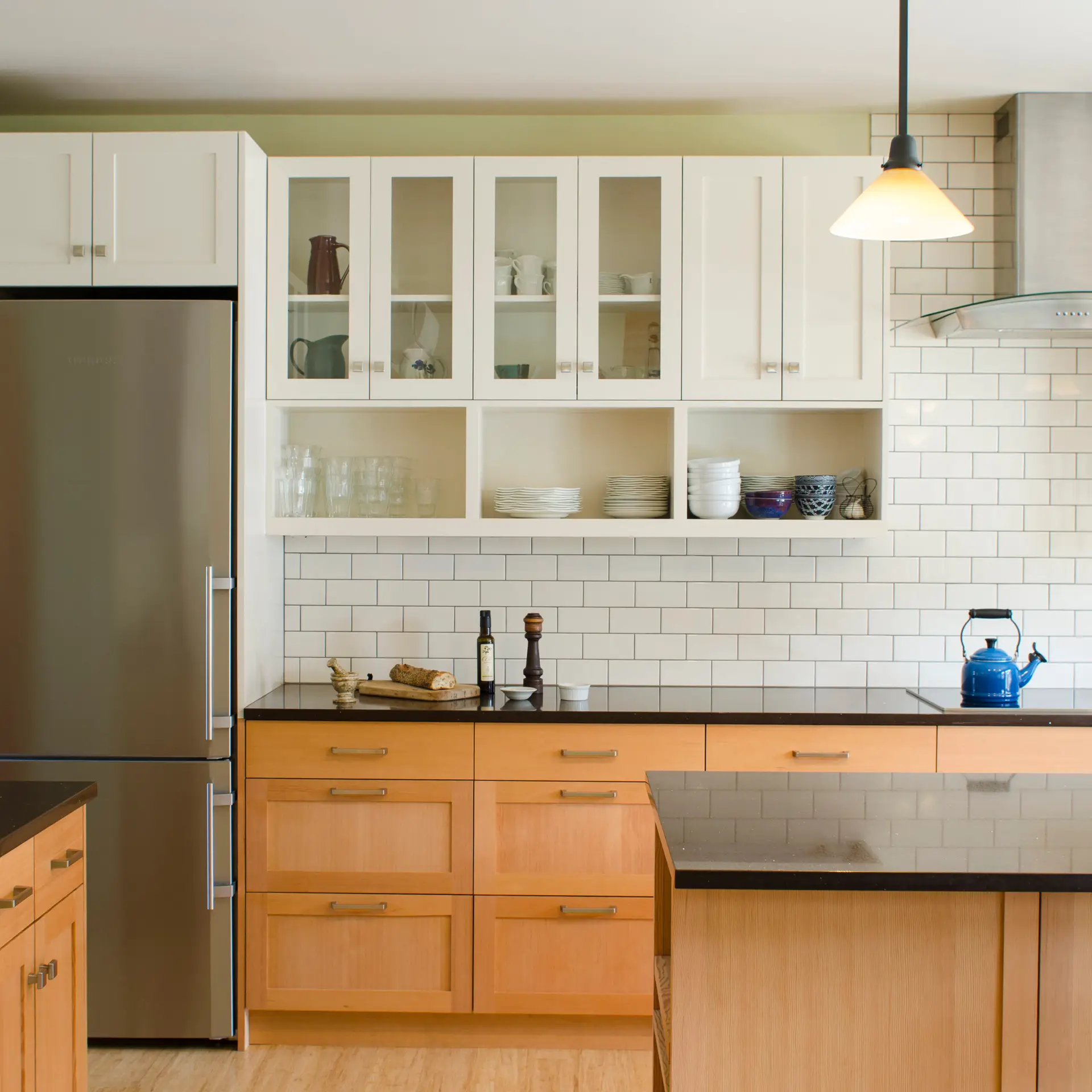 A modern, high-performance kitchen at Ankeny Row. The kitchen features a mix of white upper cabinets and warm, light-wood lower cabinets with a horizontal grain. The lower cabinets use large, deep drawers for efficient storage. A classic white subway tile backsplash with dark grout runs from the countertop to the underside of the cabinets, adding a subtle texture to the space. A black quartz countertops provides a sharp contrast to the light wood and white cabinetry.