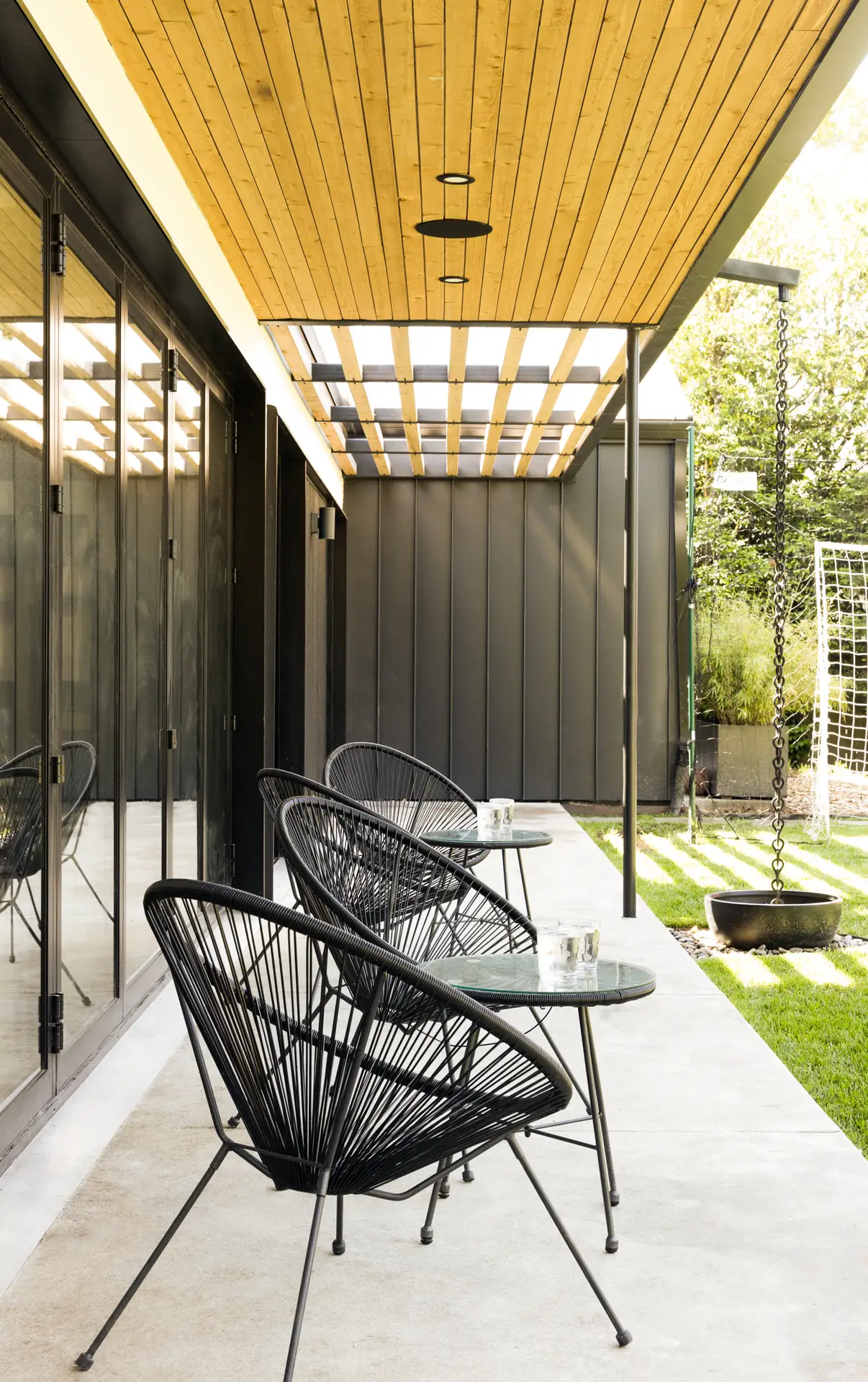 the exterior patio of the Bridlemile Residence, a LEED Platinum, zero-energy custom home in Portland, Oregon. The patio is covered by a natural wood slatted ceiling with integrated recessed lighting. A portion of the roof transitions into a wooden pergola, allowing dappled sunlight to filter through. The home's exterior is clad in a dark, vertical metal paneling that provides a sharp, modern contrast to the warm wood of the ceiling.