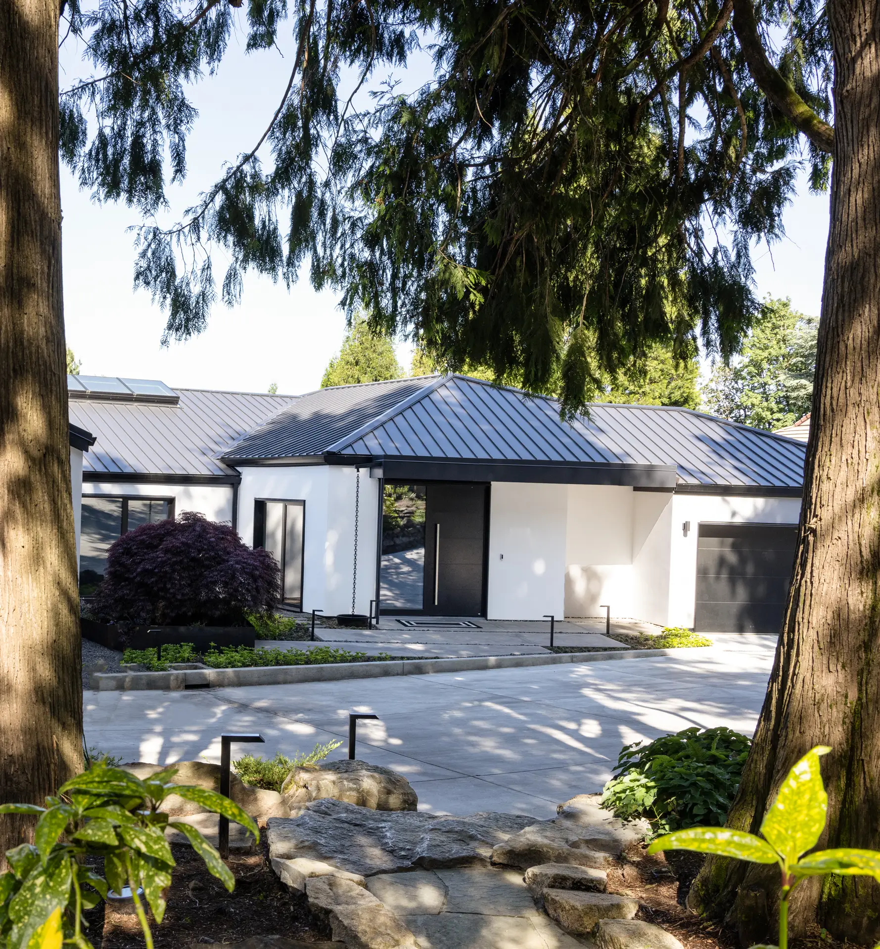 The entry, as seen through the trees, of the front of the home. Clad in white stucco, punctuated by bold black accents. The roof has a black standing-seam metal roof, which is both a modern design choice and a base for its energy-efficient solar power systems. The focal point of the facade is a large, matte black front door with a long, vertical silver handle. A matching black garage door is visible to the right.