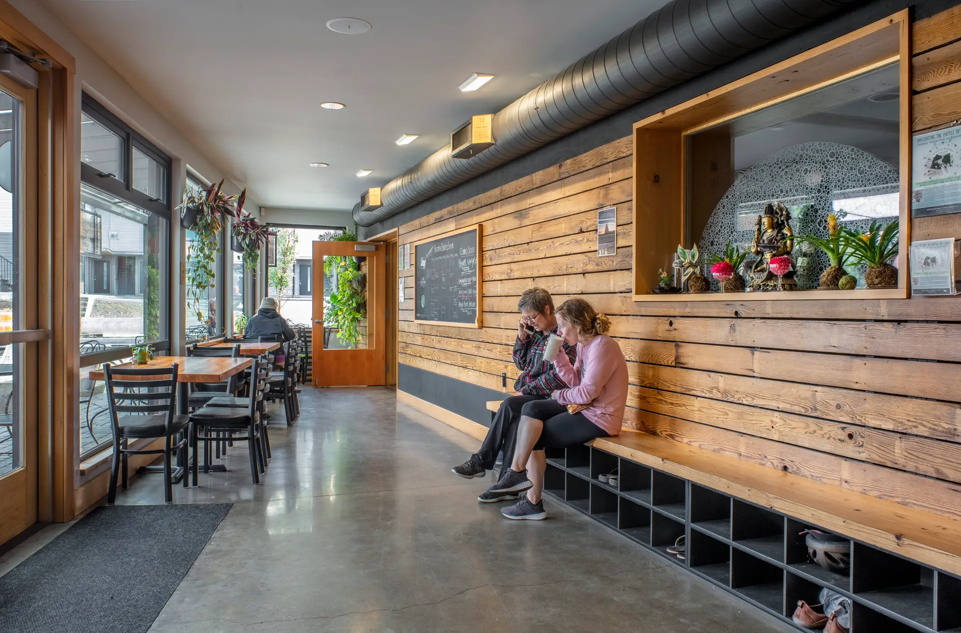 People seated at the yoga studio in The Breath Building near the entry. The entry wall features reclaimed wood and the left of the frame has tables and chairs that run along with perimeter windows of the building.