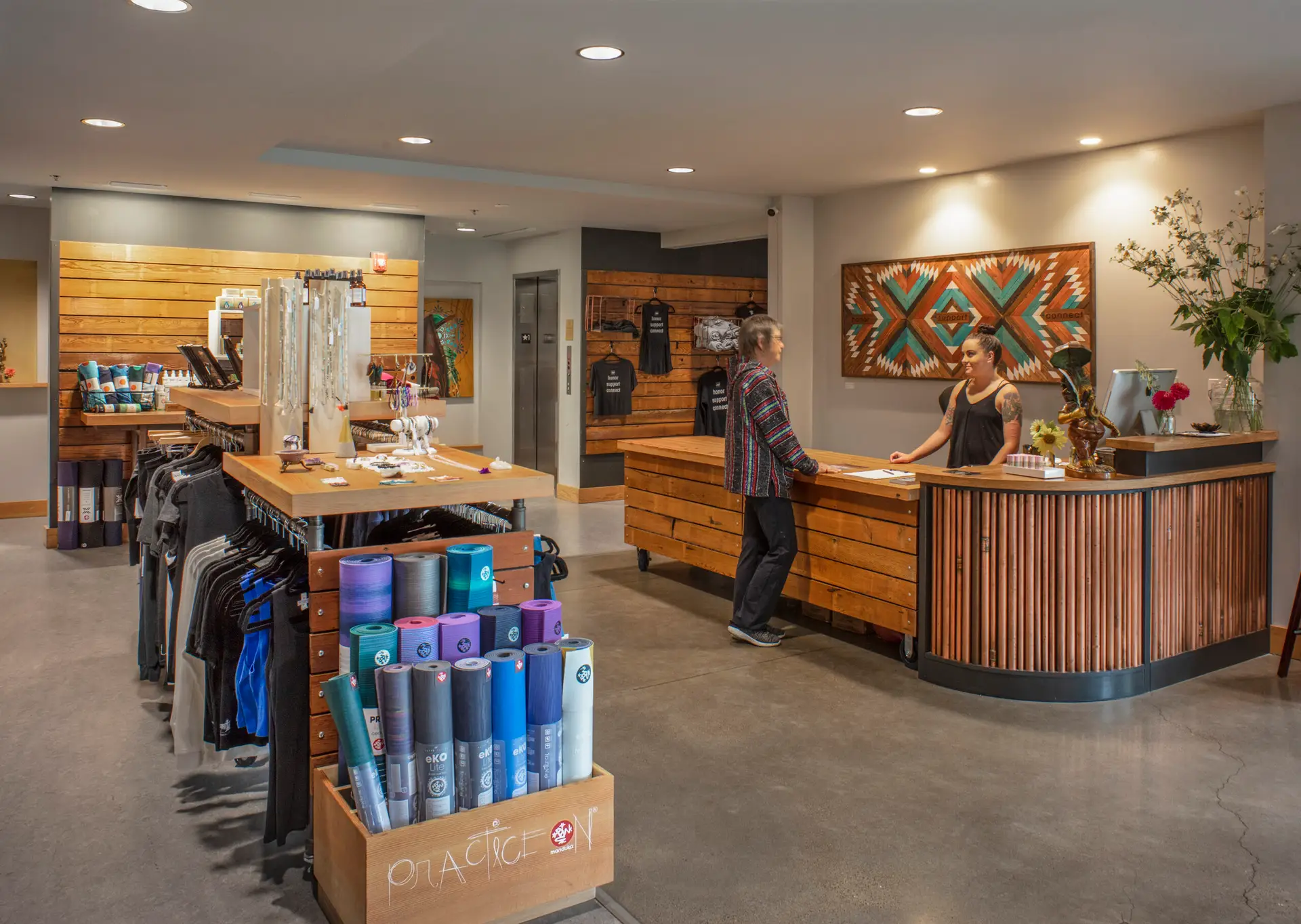 The Breath Building Yoga Studio, showing a woman at the reception desk talking to the receptionist. There are yoga mats for purchase on the let of the frame.