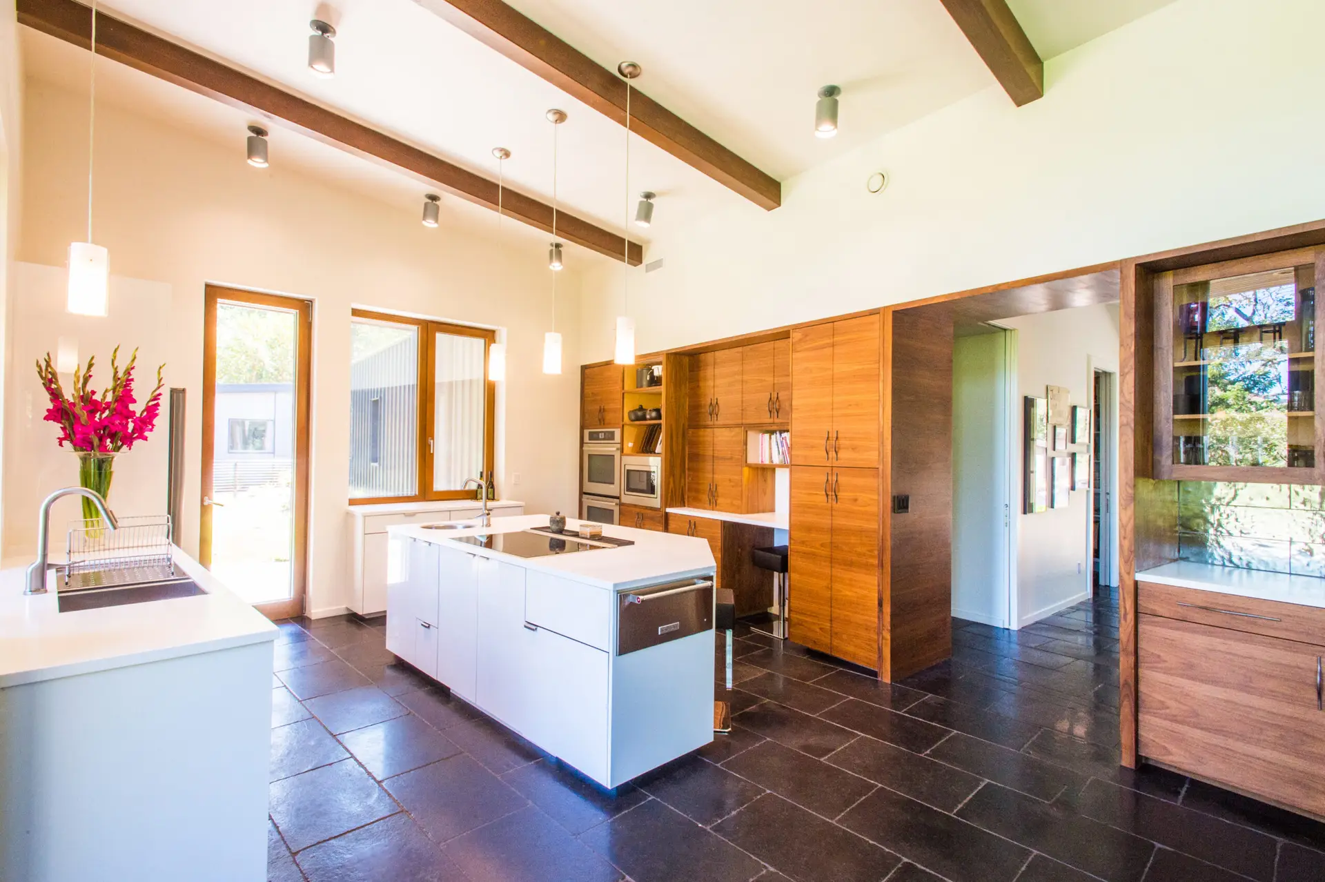 Cowhorn residence kitchen, featuring dark tile floors, a white island, and warm wood built ins. A full lite door to the exterior is to the left of frame along with a prep sink.
