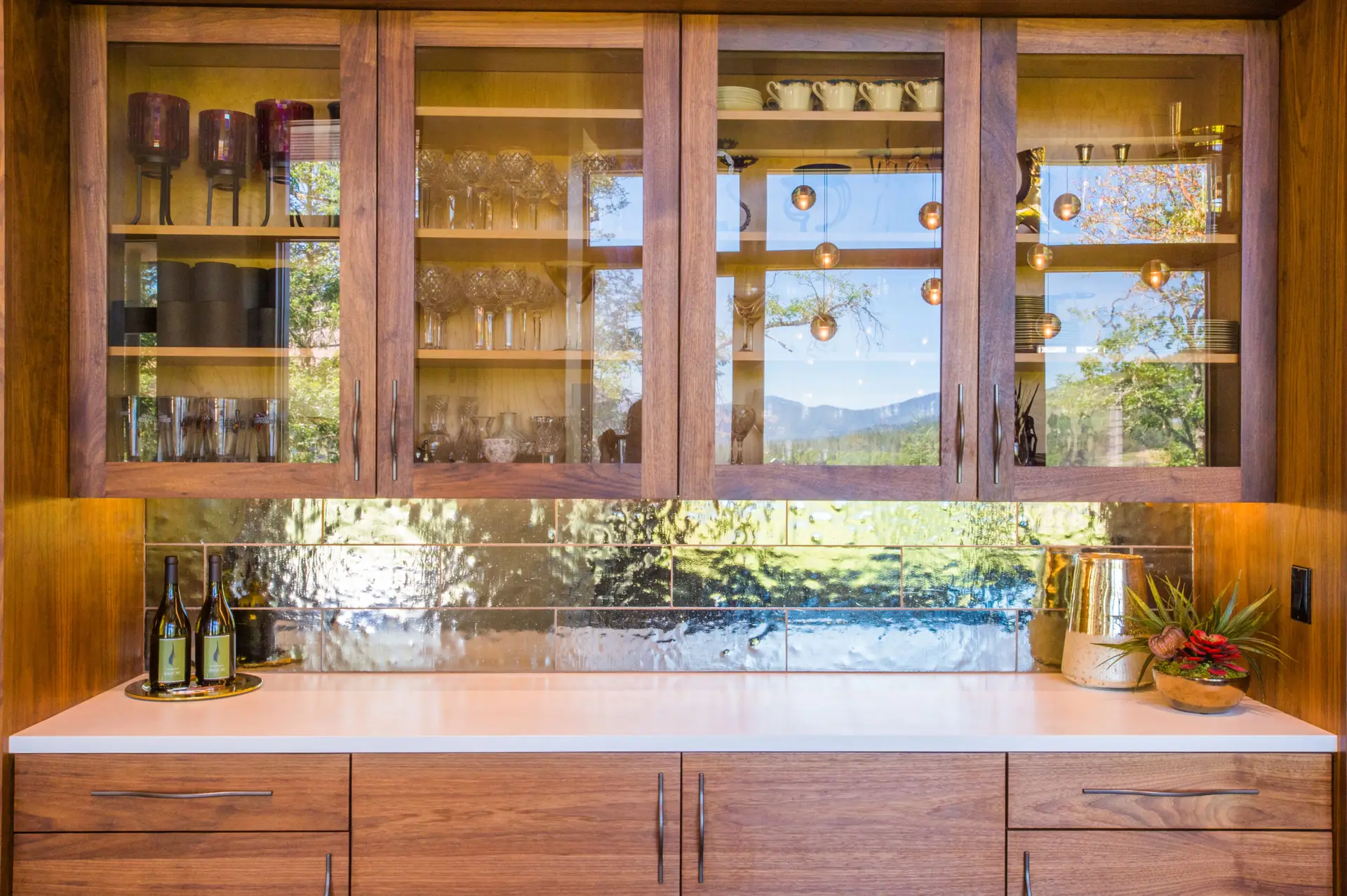 Cowhorn residence cabinets with glass upper panels and a reflective backsplash. The cabinets are a warm wood and the counter is white.