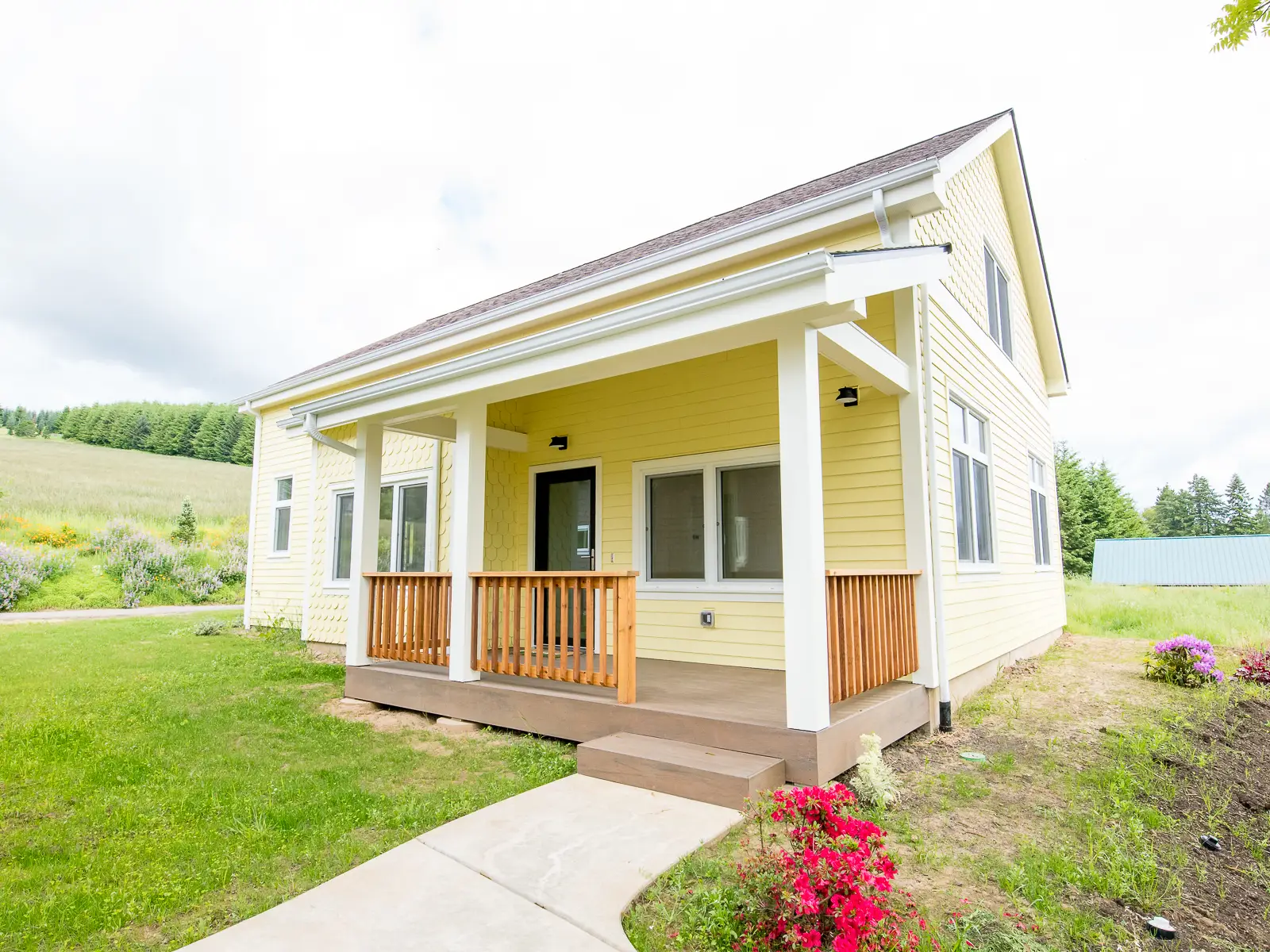 A modern farmhouse or craftsman-inspired aesthetic with a simple, two-story gabled roofline. It is clad in pale yellow horizontal lap siding, accented by white trim around the windows, doors, and porch columns. The upper gable features matching yellow shingle siding, adding texture to the design. A prominent, covered front porch serves as a central feature, supported by substantial white columns and enclosed by a natural wood railing.