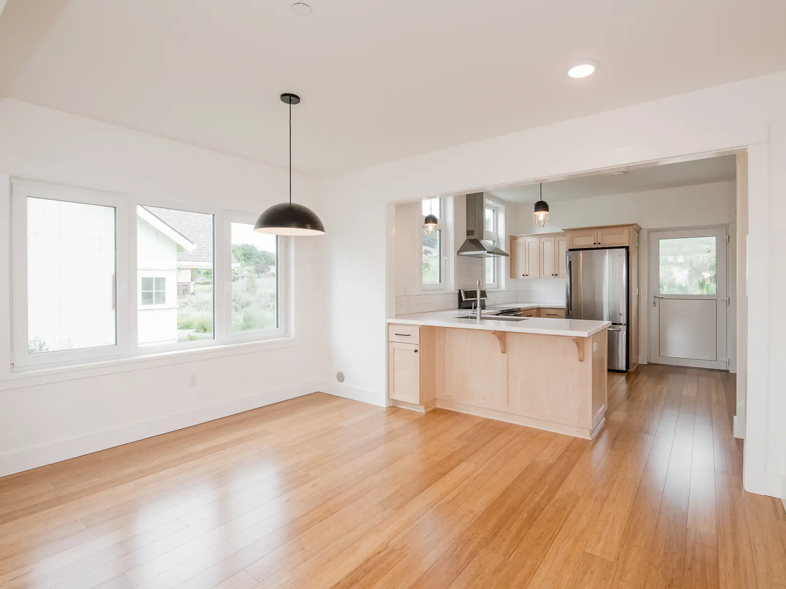 Green Grove CoHousing interior view, featuring beautiful wood floors and a light filled room from the surrounding windows. The kitchen is separated from the living by a peninsula and is highlighted by two ceiling mounted pendants.