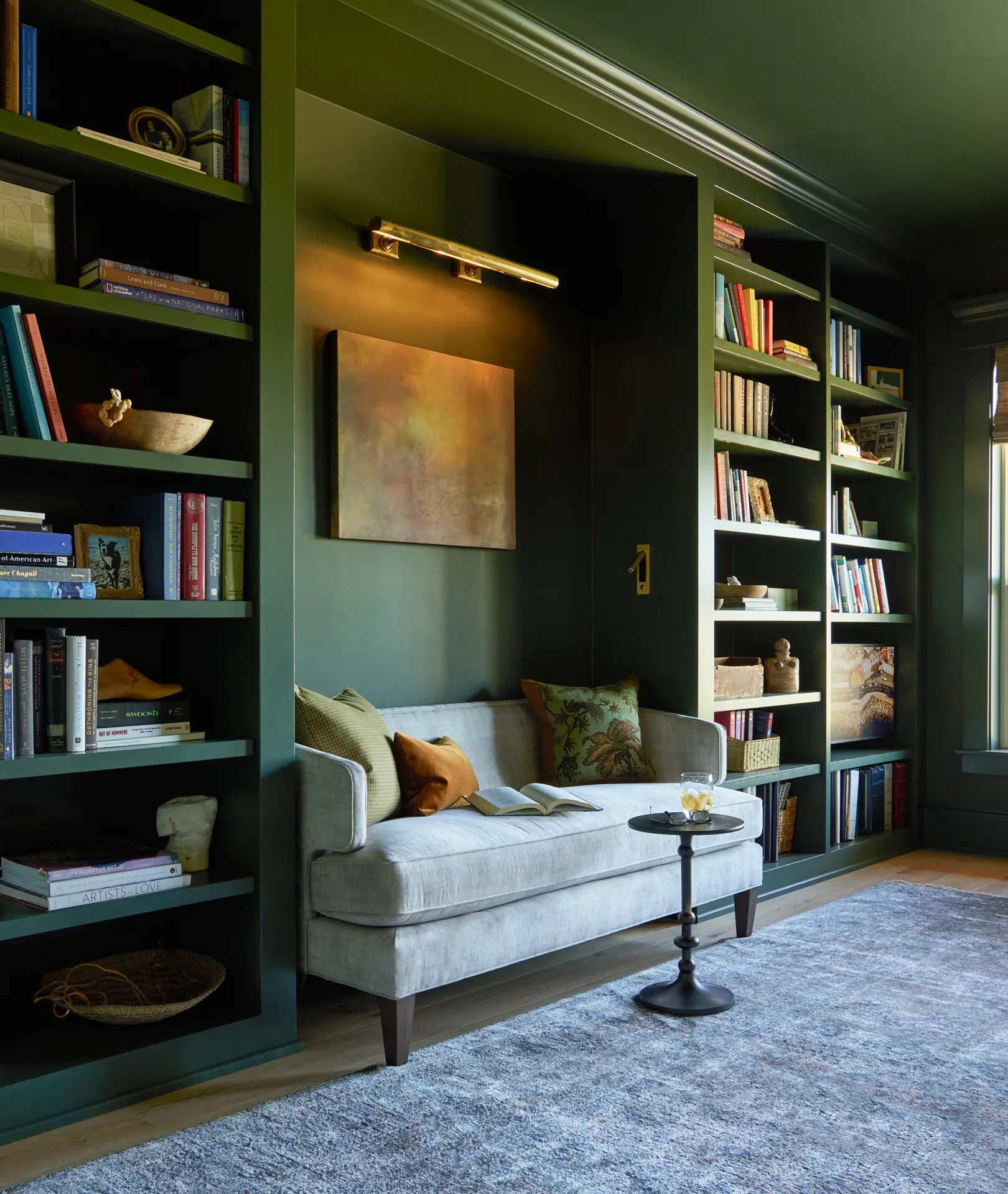 St. Paul Residence study, featuring a loveseat that is surrounded by built in bookshelves and a piece of art hung behind the loveseat. The walls and bookshelves are painted dark green.