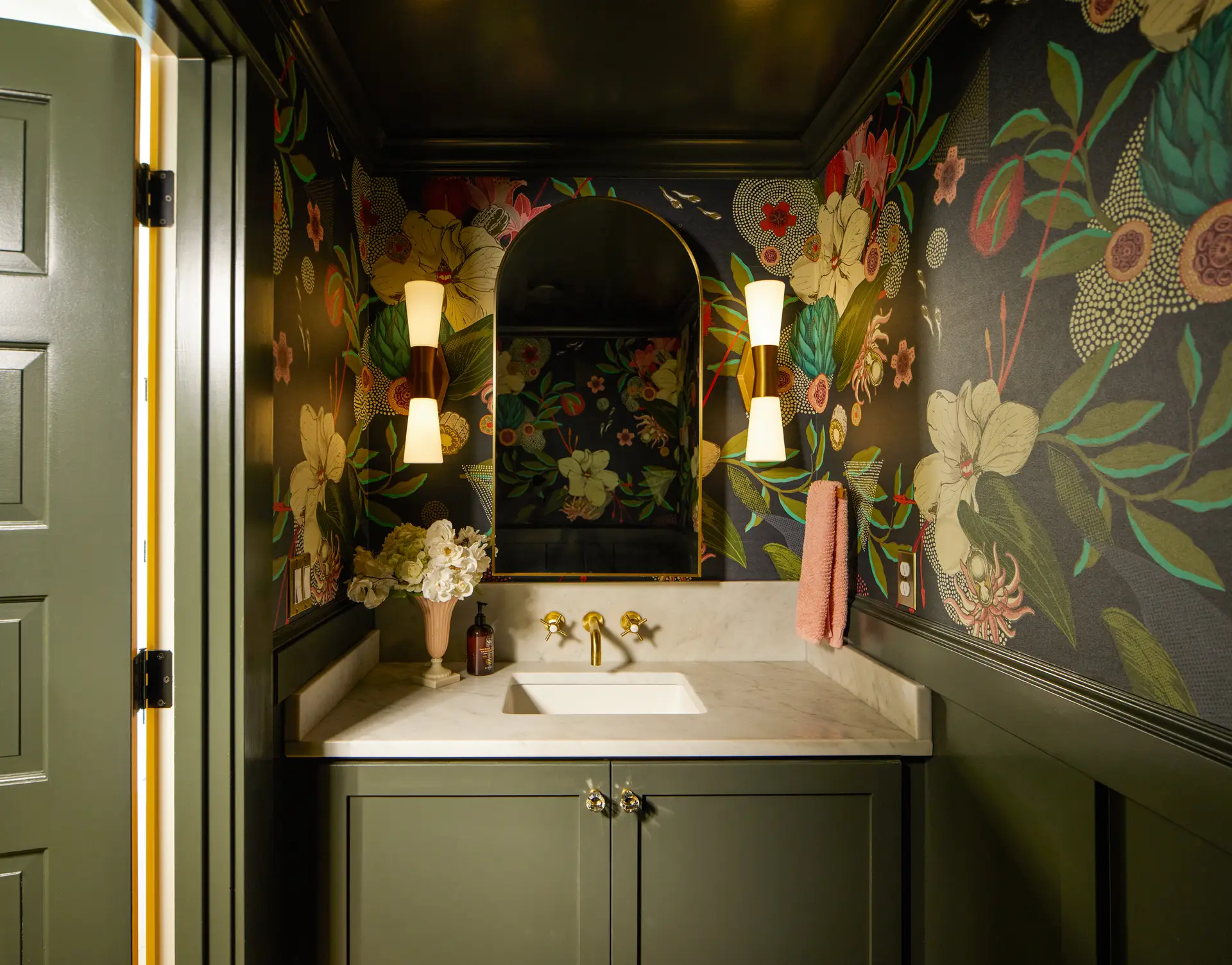 St. Paul Residence powder room, featuring a dark green and elegant design with floral wallpaper and dark green painted cabintets and lower wall panels. A curved arch mirror with flanking sconces and a wall mounted brass faucet is in the center of frame.