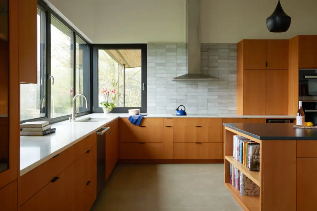 Looking at the kitchen with the sink on the left with views of the Sandy River valley, the induction cook top in the center with a stainless steel range hood. The cabintry is a warm, natural finish with minimalist black pulls. The countertops are a mix of crisp white quartz on the perimeter and a dark black quartz on the central island. The island includes integrated open shelving for a cookbook collection. A textured, light grey tile backsplash runs vertically behind the range, adding a subtle organic pattern to the space.