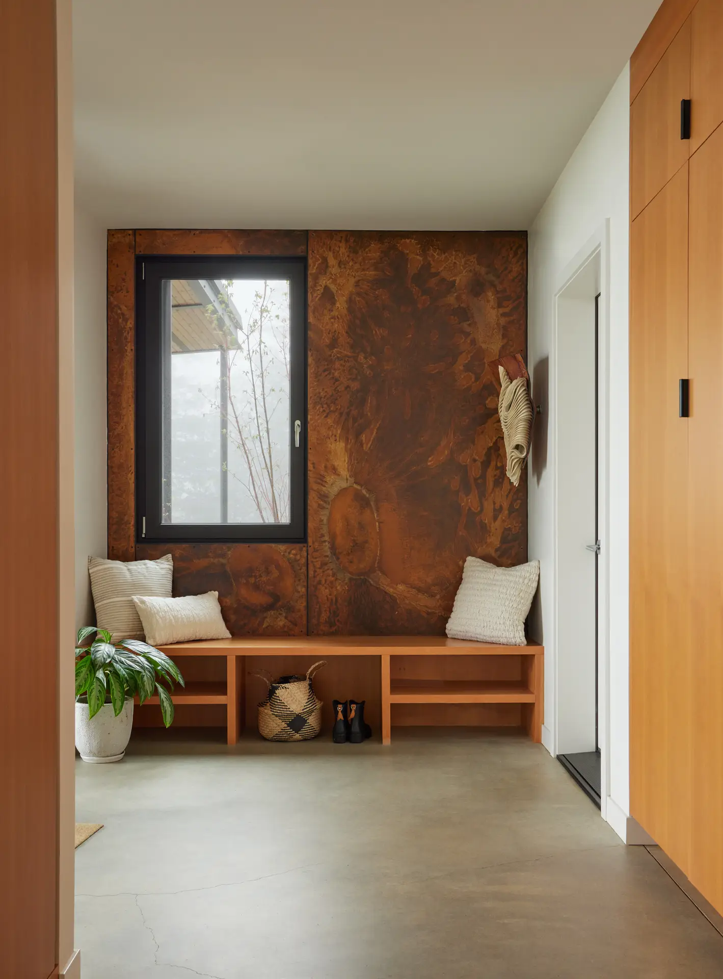 Corbett Residence entry. The focal point of the entryway is a dramatic floor-to-ceiling accent wall made of weathering steel (Corten), which intentionally patinas over time to mirror the colors of the surrounding landscape. A simple, low-profile wooden bench provides both seating and open-cubby storage for footwear. The floor is made of seamless polished concrete.