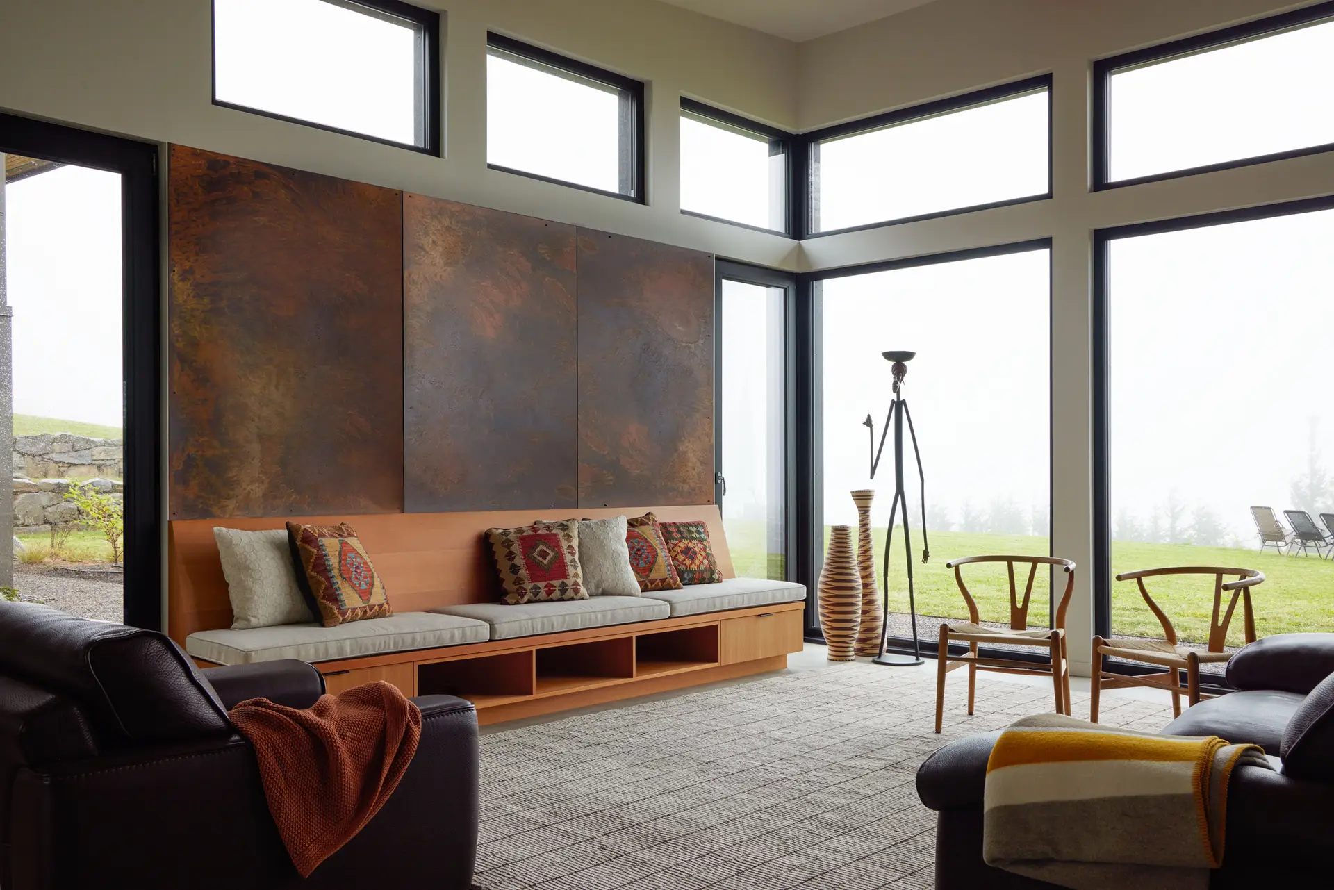 The light-filled living area of the Corbett Residence, overlooking the Sandy River through expansive floor to ceiling windows. On the left is a long, low-profile white oak bench that integrated open-cubby storage for books and accessories below. Above the couch is a three panel accent wall made of weathered steel (Corten), which introduces a rich, industrial texture that mimics the natural earth tones of the surrounding Sandy River valley.