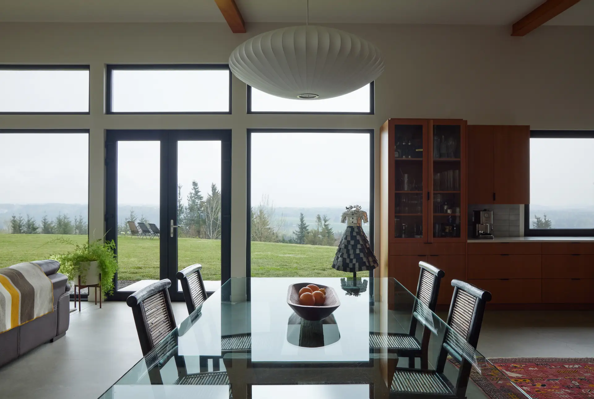 Corbett Residence diing room. This in a room with views of the Sandy River. Large black-framed triple-pane windows and a matching glass door flood the room with natural light High ceilings with wood beams flow between the adjacent kitchen and living rooms. A large, saucer-shaped Nelson Bubble Lamp hangs prominently over the dining table, serving as a mid-century modern focal point.
