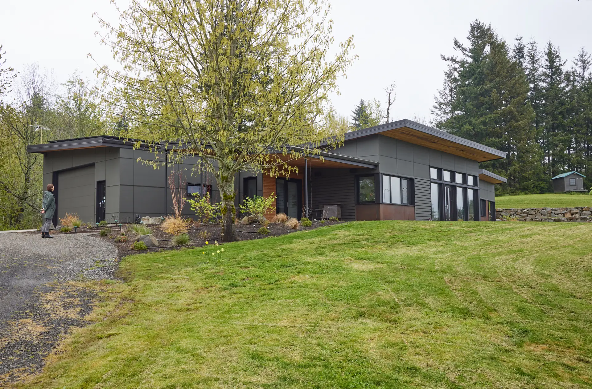 Corbett Residence, a one story, low-profile, singel-story home with shed roofs clad in a mix of dark grey fiber cement panels and warm natural wood and metal panels.
