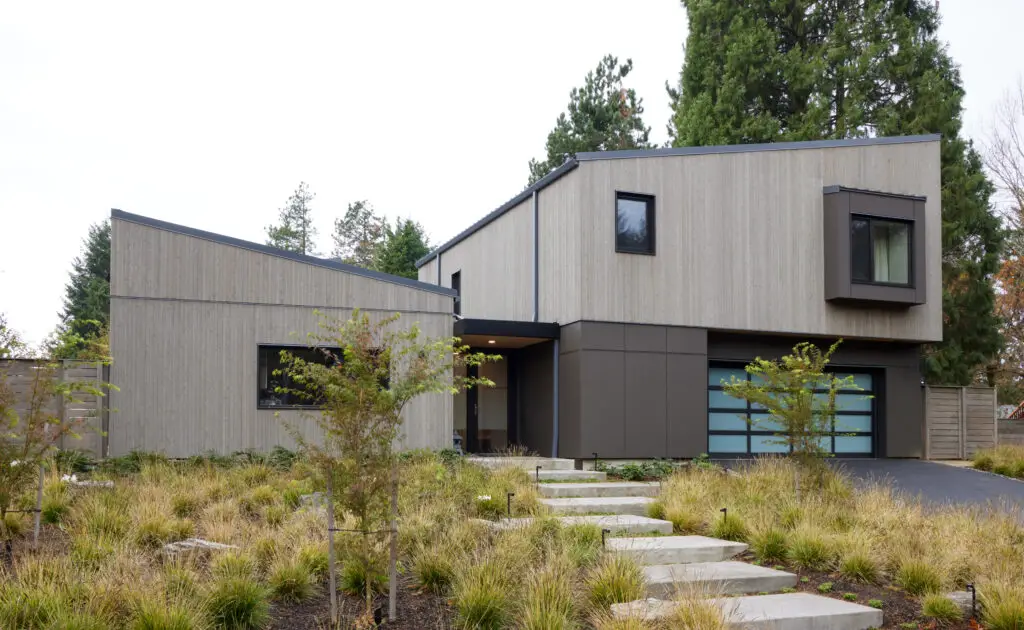 Sunset Residence view from the street, featuring sleek and modern yet inviting exterior. The building is clad with a combination of light wood siding and dark grey fiber cement siding. The landscape in front of the home is comprised of native grasses and small trees.