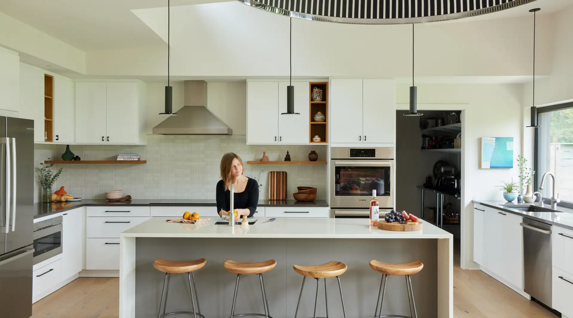 A light-filled, modern kitchen showcased in Oregon Home magazine. The space is a blend of clean, white surfaces and warm, natural wood accents. A large, white waterfall-edge island serves as the primary workspace. It features a grey base and four sculpted wood bar stools with metal legs. The kitchen uses a mix of crisp white upper and lower cabinets with minimalist black hardware. Open wood-lined cubbies and floating wood shelves break up the white cabinetry, adding warmth and display space for ceramics.