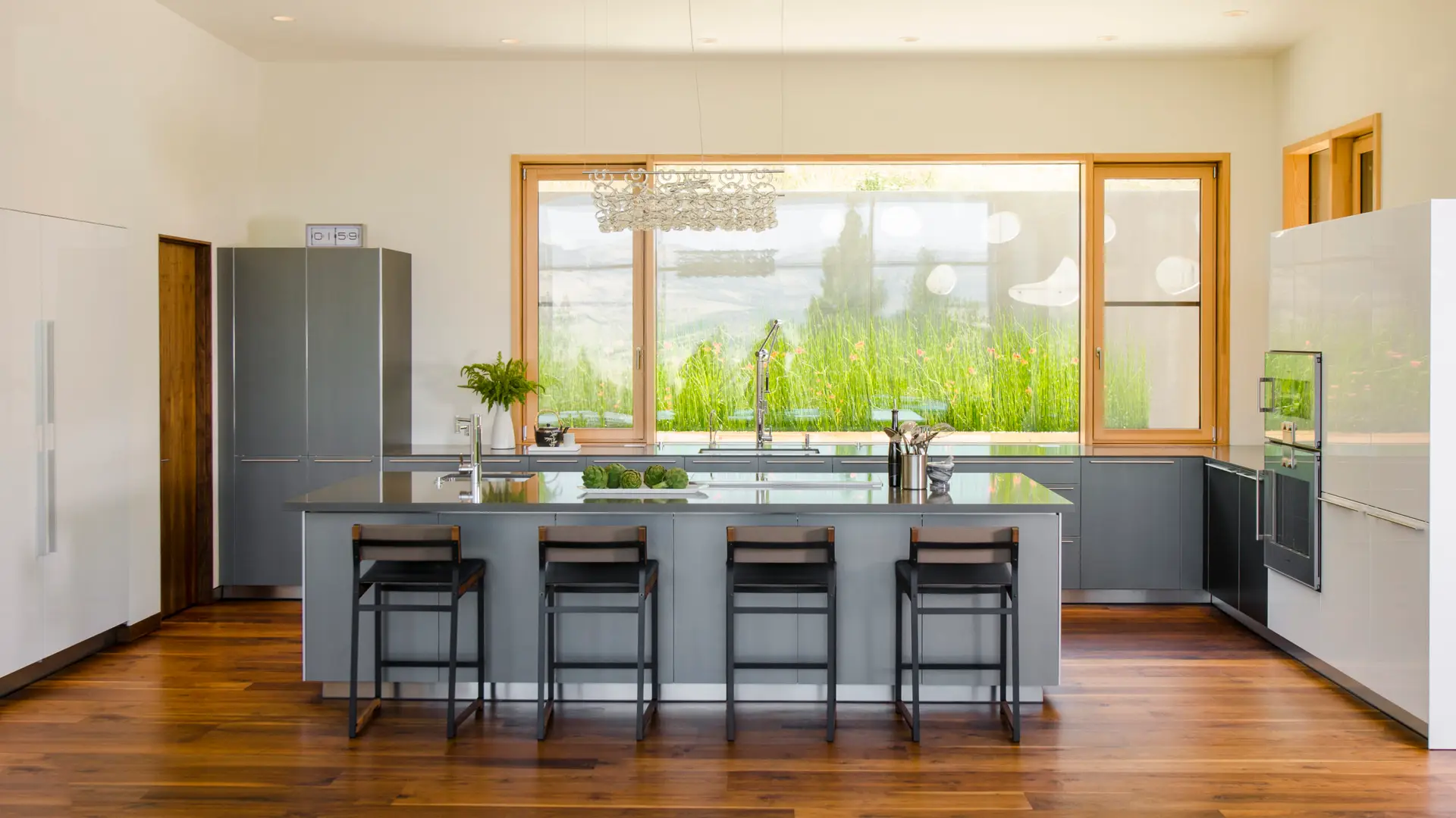 Fineline House is characterized by a "quiet modern" aesthetic that emphasizes natural light and healthy, high-quality materials. A monolithic grey island serves as the primary workspace and gathering hub, featuring an undermount sink and seating for four contemporary stools with dark wood backrests. The focal point is a massive, wood-framed triple-pane window, which provides a panoramic view of the lush, green hillside and valley while ensuring superior thermal insulation