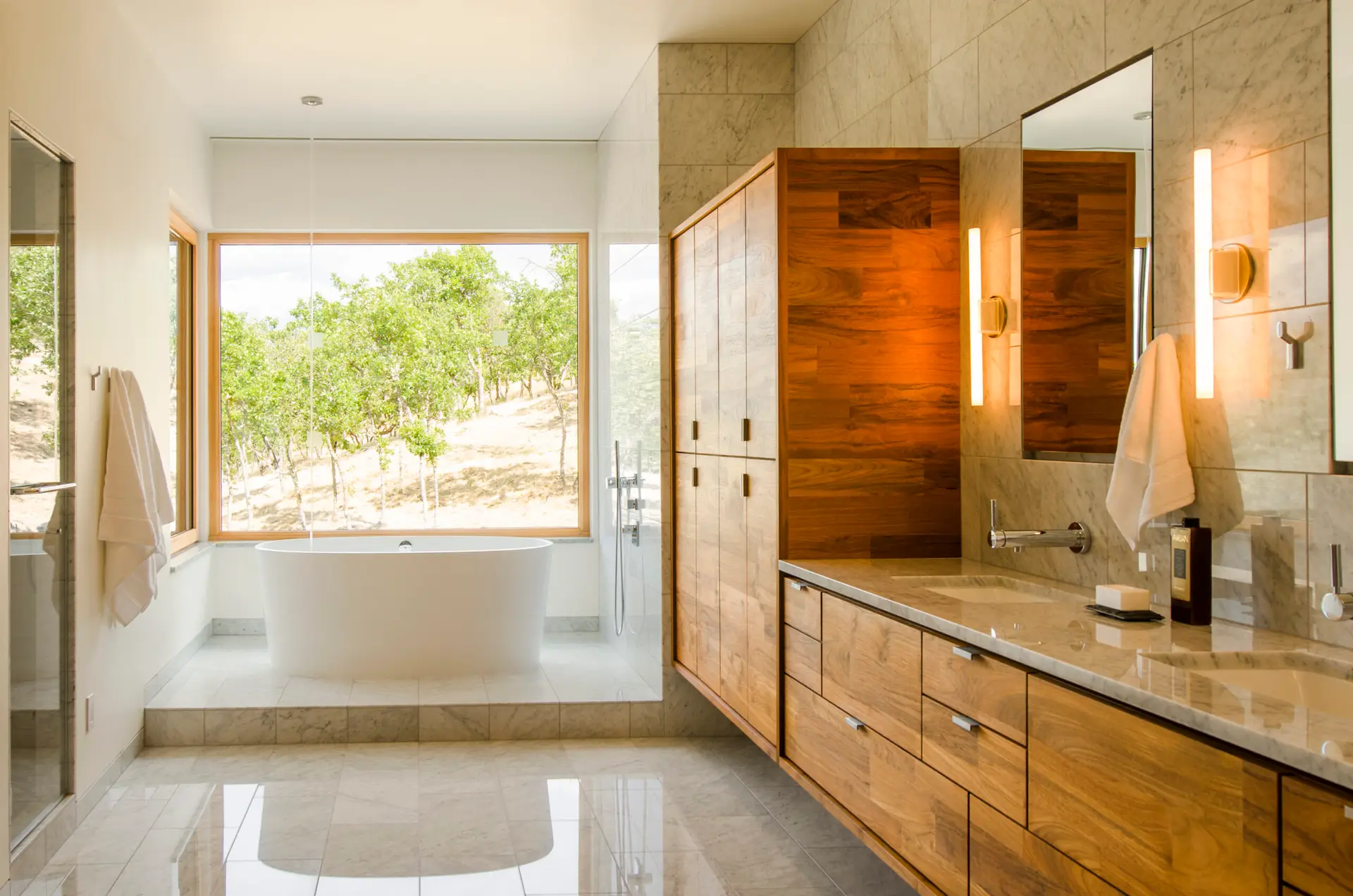 Fineline Custom Home primary bathroom featuring a deep, white freestanding oval bathtub positioned on a raised marble-tiled platform and offers a panoramic view of the surrounding forested hillside. On the right is a large, floating walnut vanity and matching floor-to-ceiling linen cabinet. The vanity is topped with a grey quartz countertop featuring two rectangular undermount sinks and sleek, chrome wall-mounted faucets.