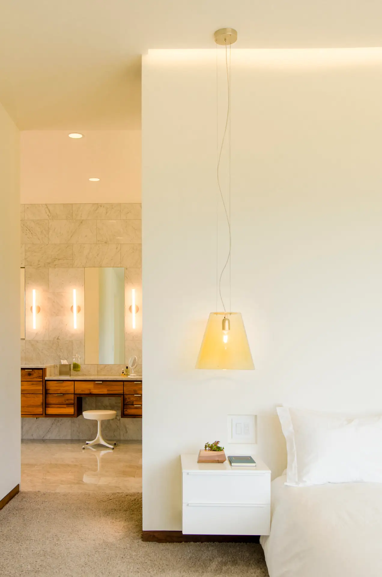 Fineline Custom Home primary bedroom looking into the luxury bathroom. A floating white nightstand that features a clean, handle-less design. Above the nightstand hangs a translucent, amber-colored tapered glass pendant light, providing warm, focused task lighting. The bathroom is finished with large-format marble tiles on the floors and walls. It features a custom floating walnut vanity and a dedicated makeup station with a white pedestal stool.
