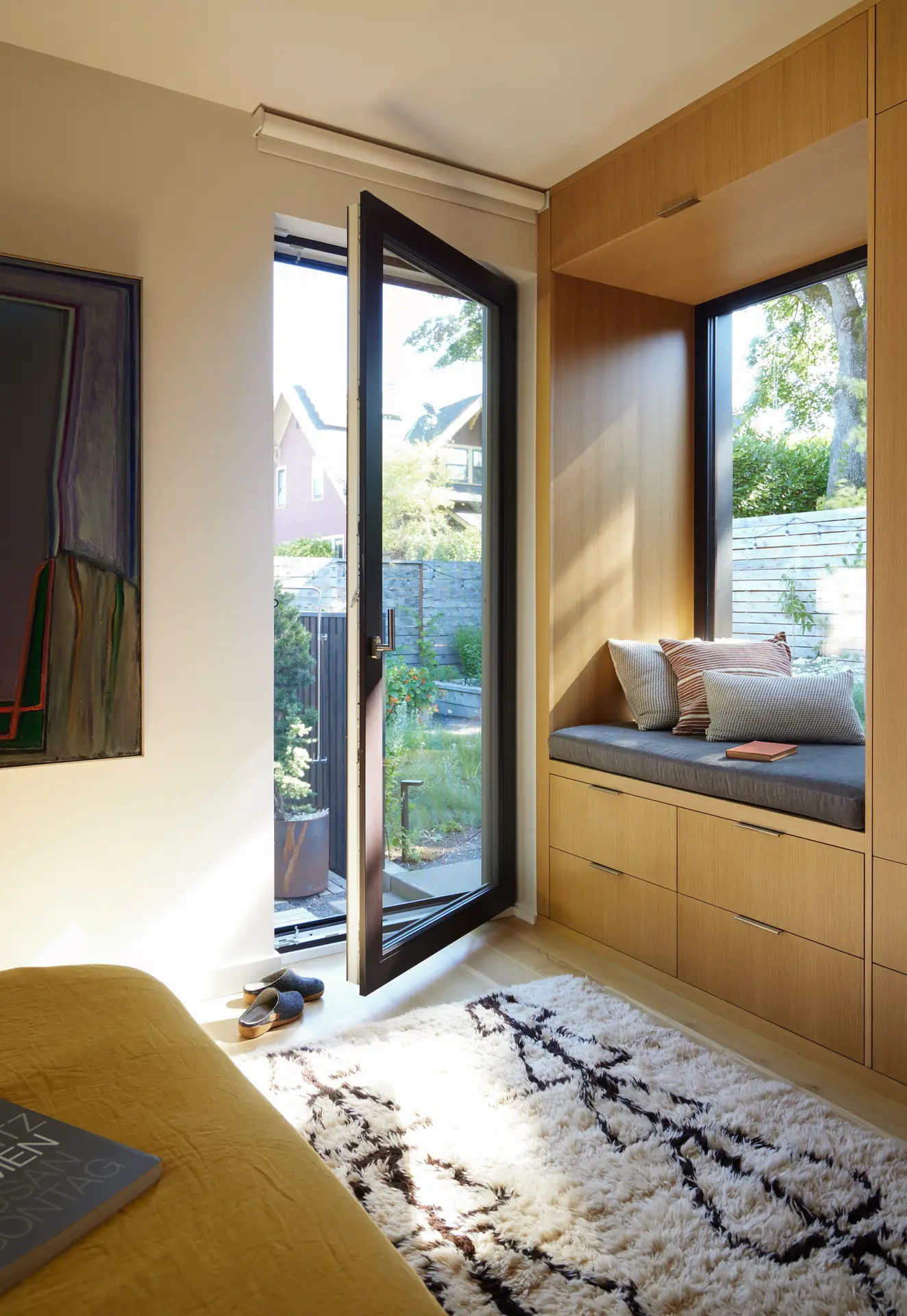 the Richmond residence bedroom, featuring a view of the full glass door that accesses the backyard. A built in window seat with drawers below is on the right side of the frame and the bed is in the foreground. A high pile rug leads out to the glass door.