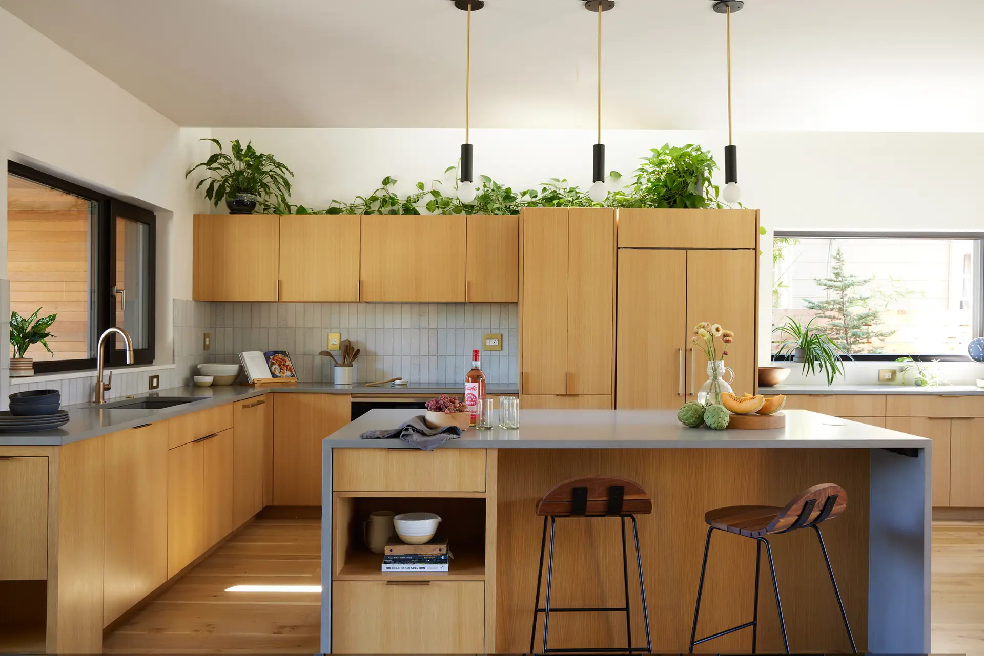 the Richmond residence kitchen, highlighting the wood cabinets, light blue tile backsplash, clerestory windows, and plats on top of the upper cabinets. The foreground shows an island with waterfall countertop and 2 counter height stools.