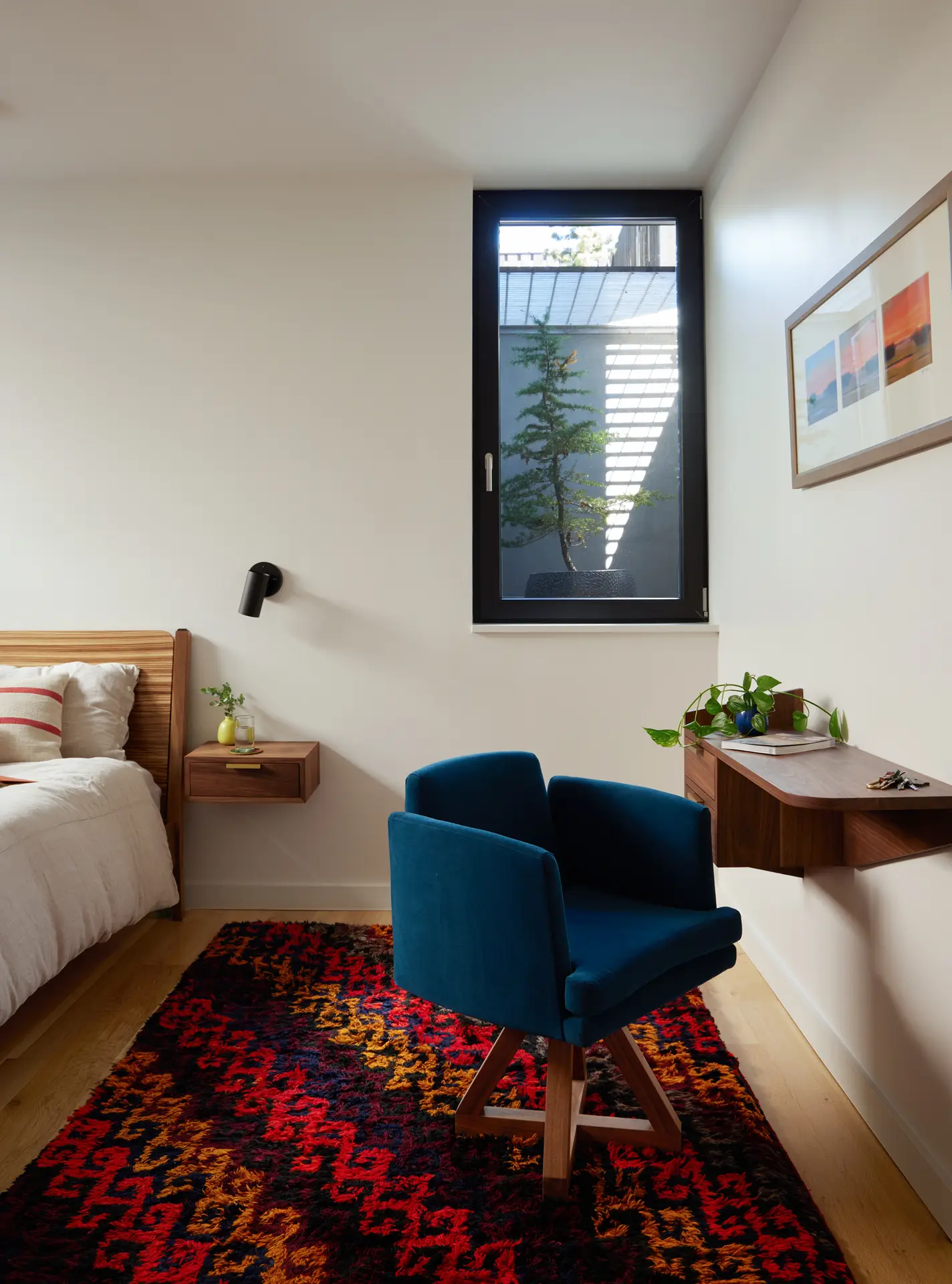 a view of a bedroom at Richmond residence. In the foreground is a beautiful red, yellow and maroon rug with a blue office chair and a built in desk. To the left of the frame is the bed. A tilt/turn window is in the center right of the frame with a view out.