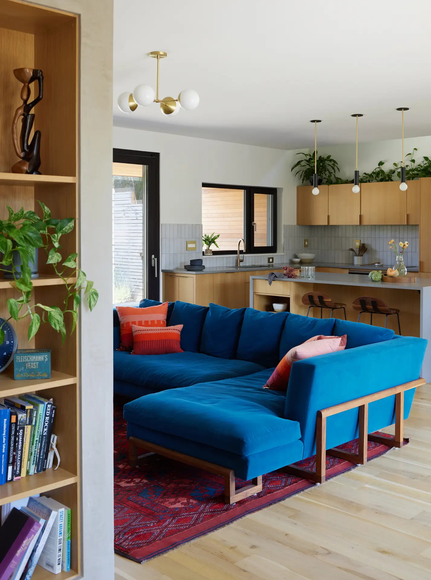 the living room at Richmond residence, showing a blue couch with orange accent pillows. Beyond is the kitchen which features light wood cabinets. In the foreground is a built in bookshelf
