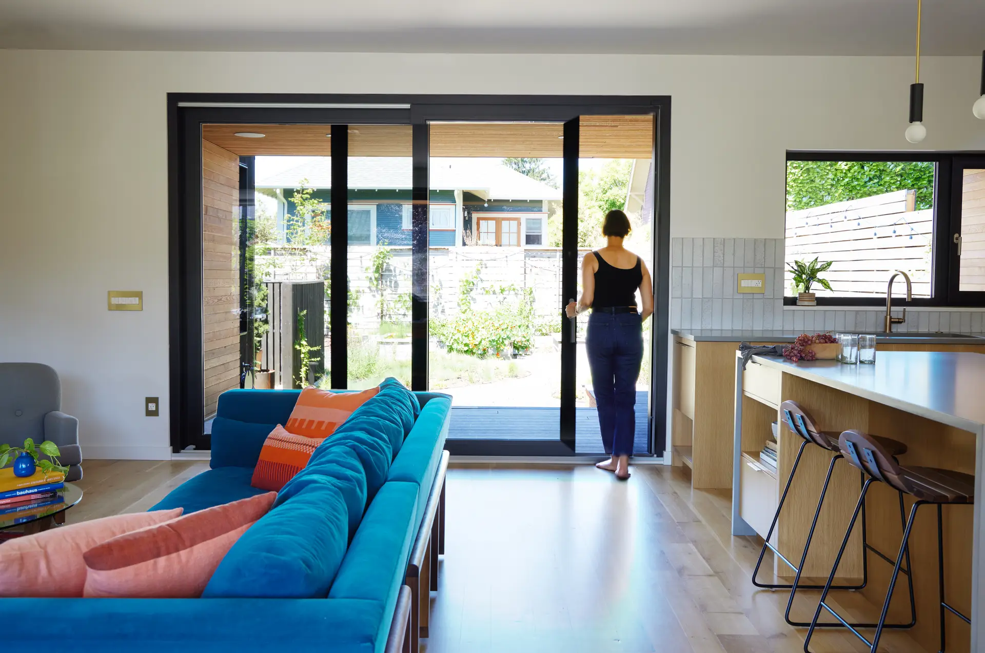 the living room at Richmond residence, showing a blue couch with orange accent pillows. Beyond is a large sliding glass door with a woman walking outside