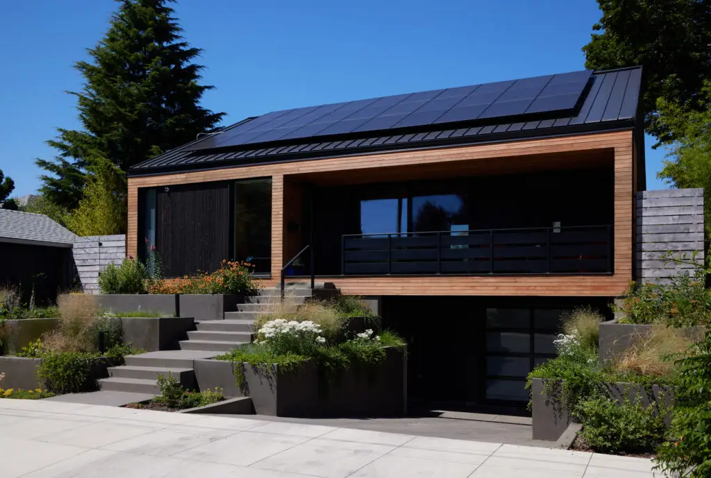 Richmond residence's front entry with stairs leading up to the front porch. The building highlights a combination of light wood siding and darker roof and complimentary siding. A large photovoltaic array is visible from this view of the roof.