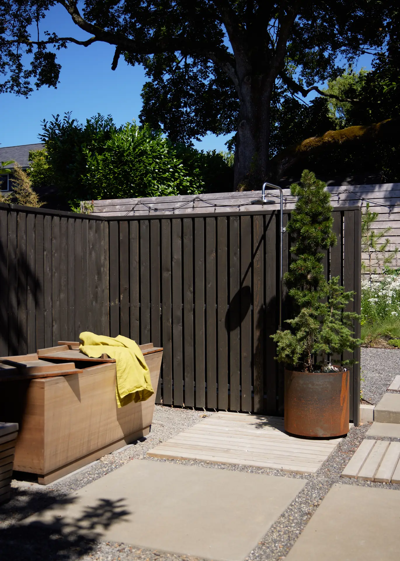 the exterior patio at Richmond residence. An outdoor shower with a medium size potted tree is shown alongside a dark wood fence.
