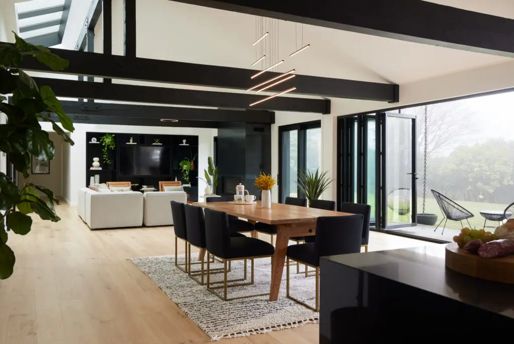 Open-concept great room of the Bridlemile Residence. The room has vaulted ceilings with exposed heavy-timber trusses that have been painted black for a dramatic effect. A 15-foot-long triple-pane skylight and a 22-foot-long accordion door flood the space with natural light while providing seamless indoor-outdoor access to the expansive yard. At the far end of the living area is a custom steel fireplace and a large black built-in media wall that contrasts with the bright white walls and light-wood flooring.