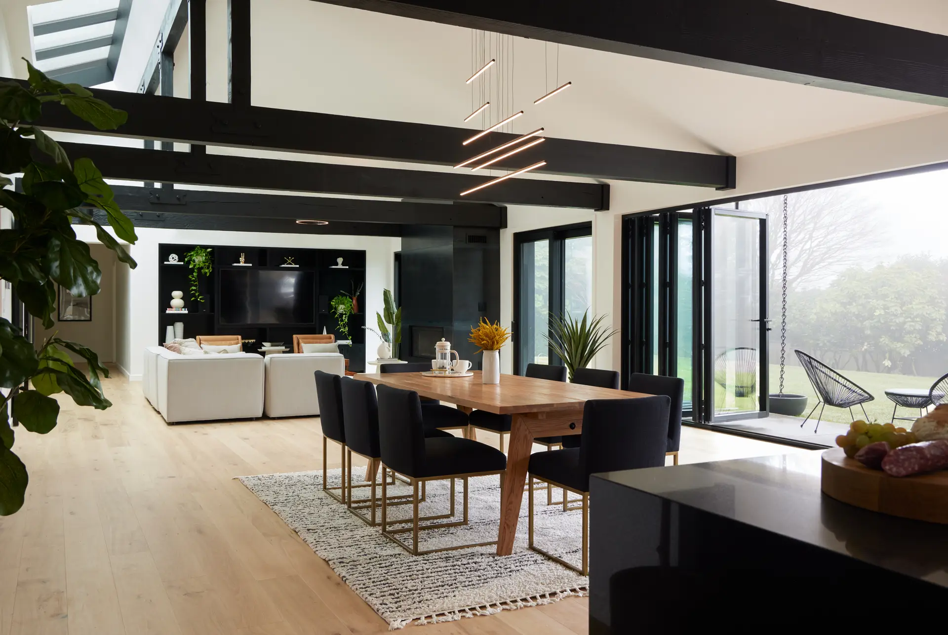 Open-concept great room of the Bridlemile Residence. The room has vaulted ceilings with exposed heavy-timber trusses that have been painted black for a dramatic effect. A 15-foot-long triple-pane skylight and a 22-foot-long accordion door flood the space with natural light while providing seamless indoor-outdoor access to the expansive yard. At the far end of the living area is a custom steel fireplace and a large black built-in media wall that contrasts with the bright white walls and light-wood flooring.