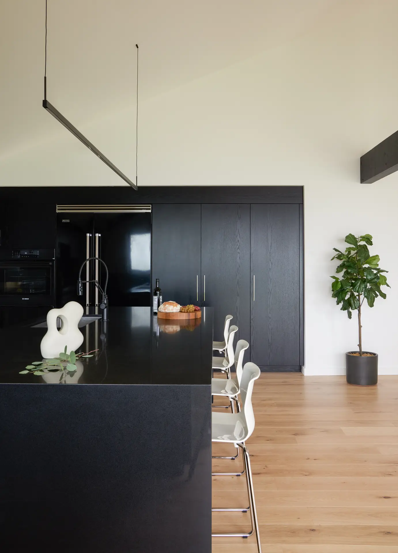 Modern kitchen of the Bridlemile Residence he kitchen features a bold, high-contrast palette that defines the home's "modern craftsman" aesthetic. Full-height, custom matte black cabinetry with a vertical wood grain provides extensive storage and a dramatic backdrop. A large, monolithic island is topped with recycled black quartz countertops, serving as a functional workspace and social hub. Wide-plank white oak flooring adds organic warmth and balances the dark architectural elements.