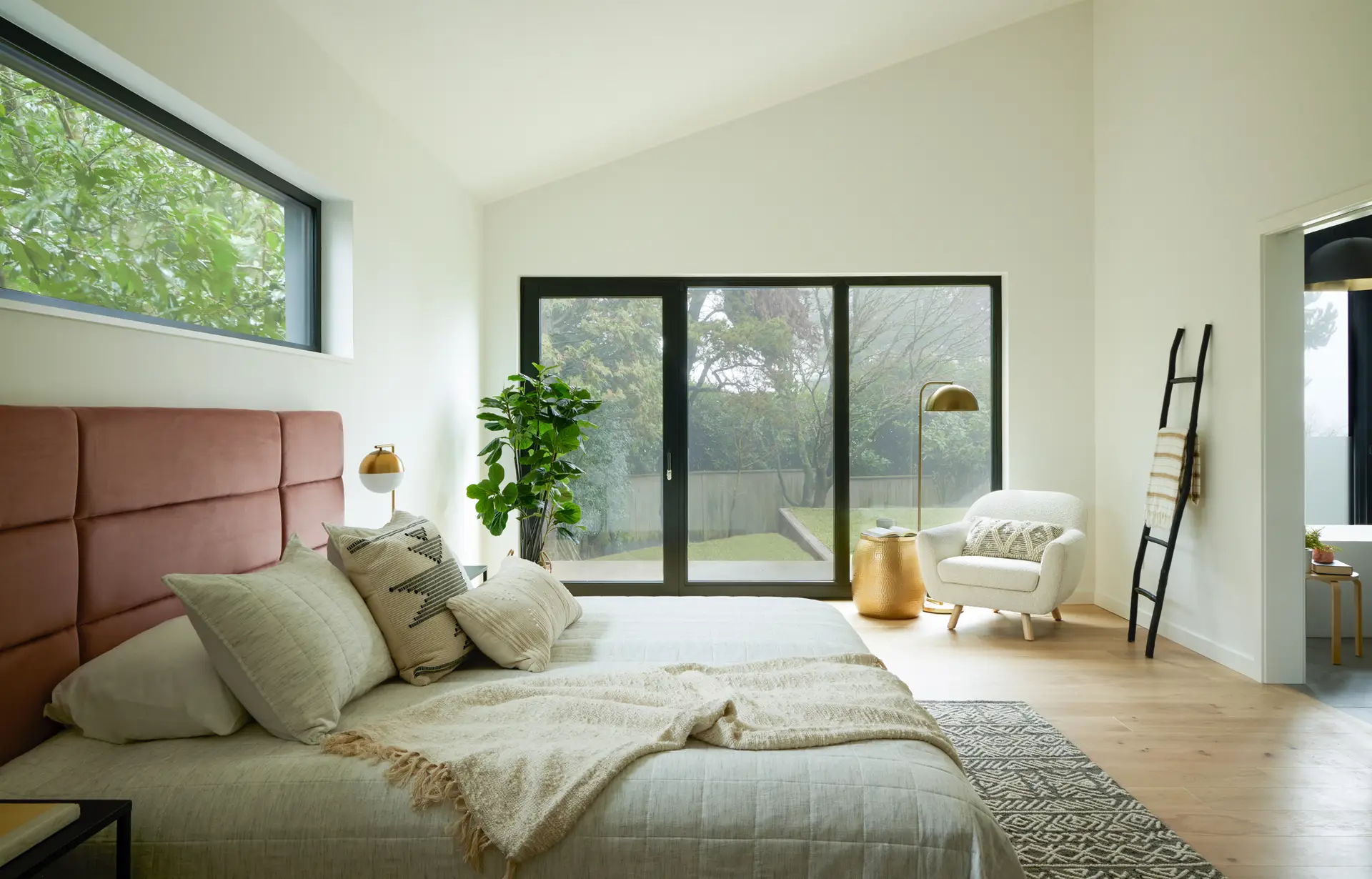 The primary bedroom of the Bridlemile Residence. High, sloping ceilings enhance the room's volume and airy feel. Large black-framed triple-pane windows and a sliding glass door provide abundant natural light and forest views while maintaining a quiet, energy-efficient environment. The sliding door opens directly to the backyard, seamlessly connecting the sleeping area with the lush outdoor landscape