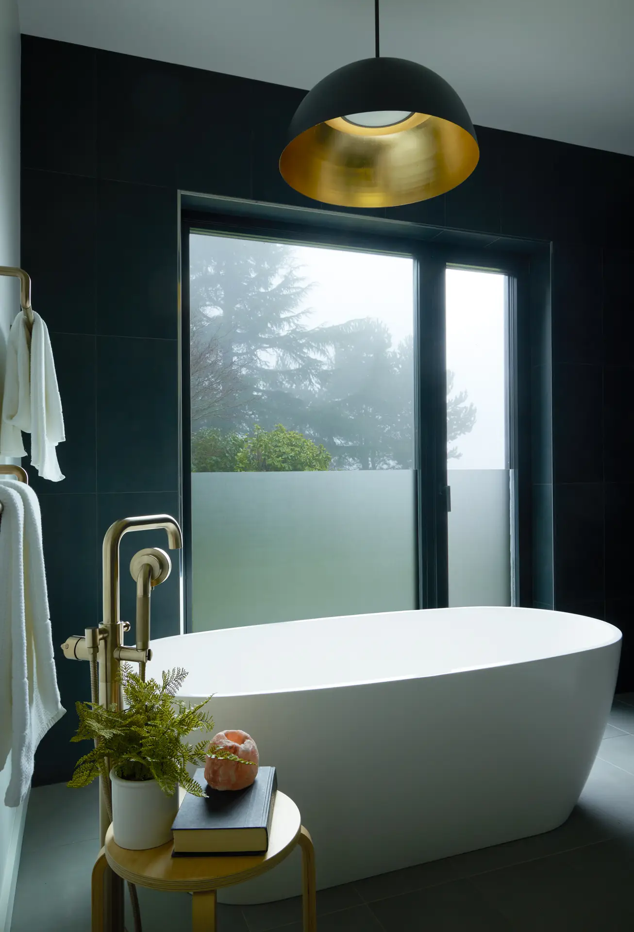 The primary bathroom of the Bridlemile Residence. A deep, white freestanding oval bathtub serves as the central focal point, positioned directly in front of a large window. The walls are clad in large-format dark charcoal tiles, providing a bold contrast to the bright white tub and the light wood of the accent furniture. A prominent brushed-gold floor-mounted tub filler adds a touch of refined elegance and warmth to the cool-toned space.