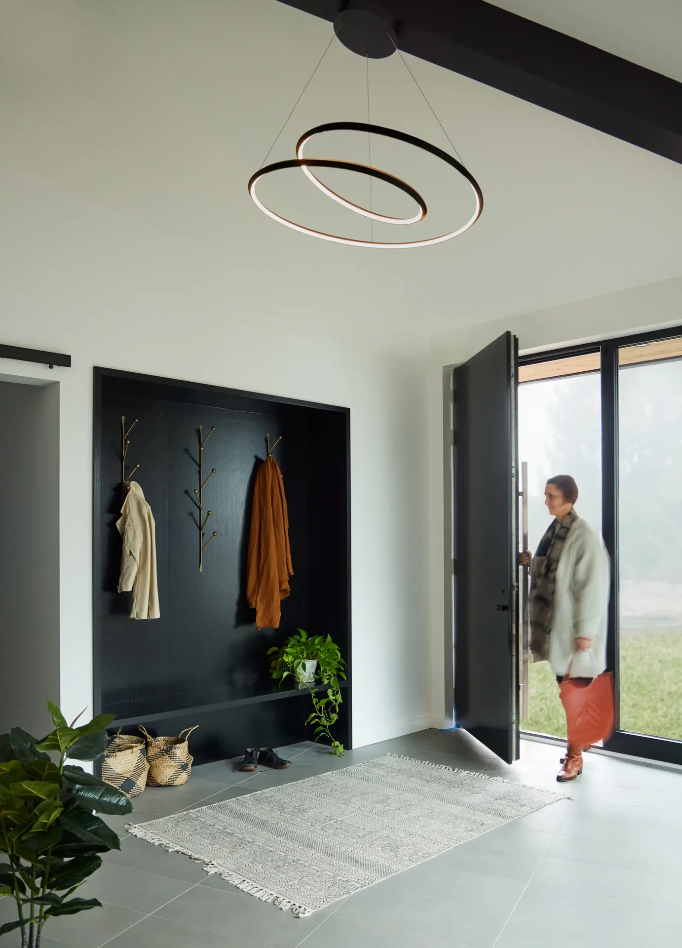 The airy, modern entryway of the Bridlemile Residence. A large, matte black front door with a long silver handle opens to a light-filled space. To the left, a recessed black mudroom niche features built-in seating, a shelf for shoes, and gold tree-branch-style coat hooks. A striking black halo-style LED pendant light with two interlocking rings hangs from the vaulted ceiling, serving as a contemporary focal point. A prominent black-painted heavy-timber beam spans the ceiling, adding a structural and dramatic element to the white-walled interior.