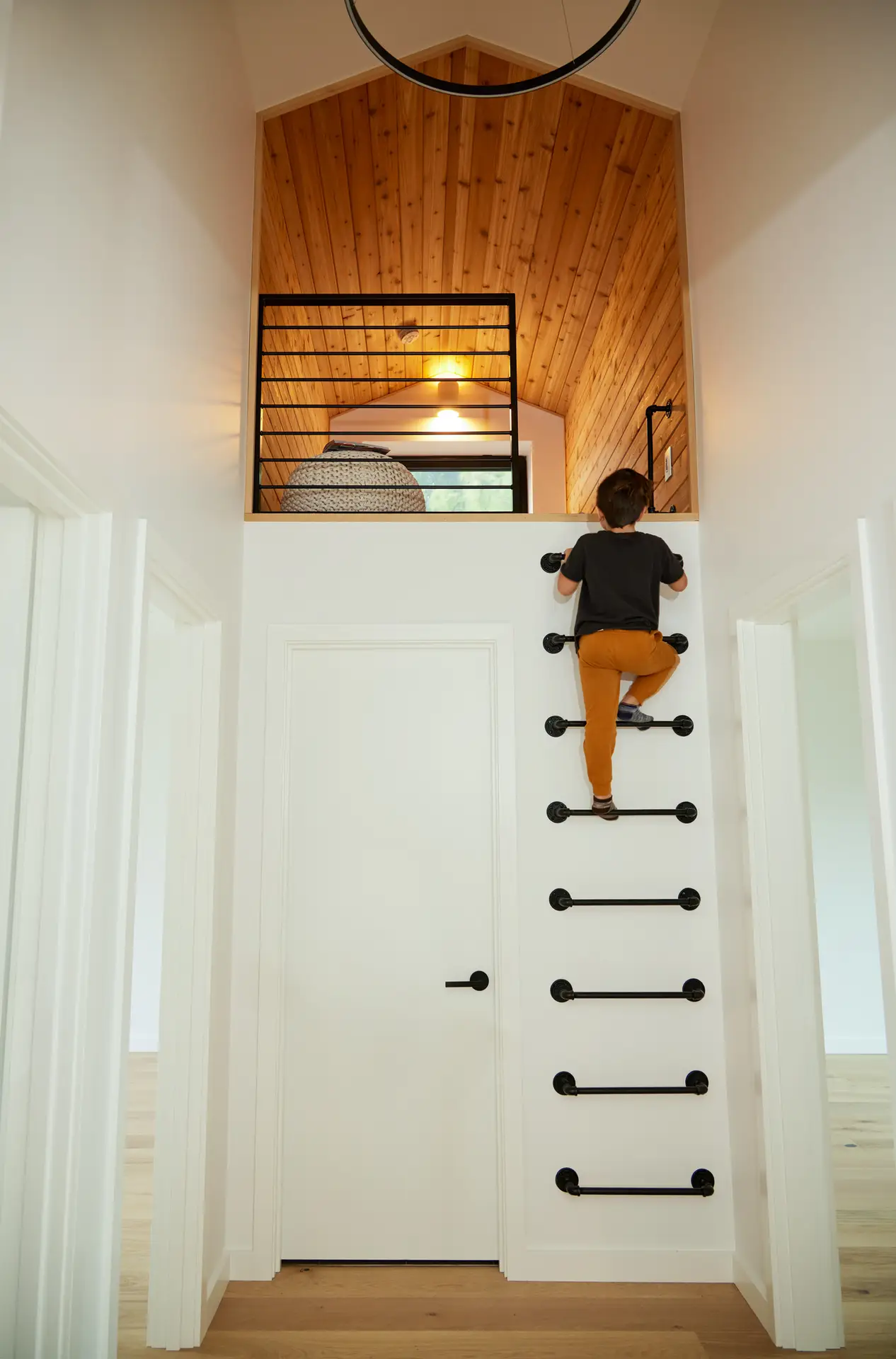 A hallway leading to a reading loft with a built-in climbing ladder. A series of black, industrial-style pipe rungs are mounted directly to the white wall, serving as a permanent ladder. The destination is a small loft nestled under a gabled roof. It is entirely clad in warm natural wood paneling on the walls and ceiling, creating a distinct, "nest-like" contrast to the crisp white hallway below. A black horizontal-slat metal railing provides safety for the loft, while a warm overhead light and a small window ensure the space is bright and inviting.
