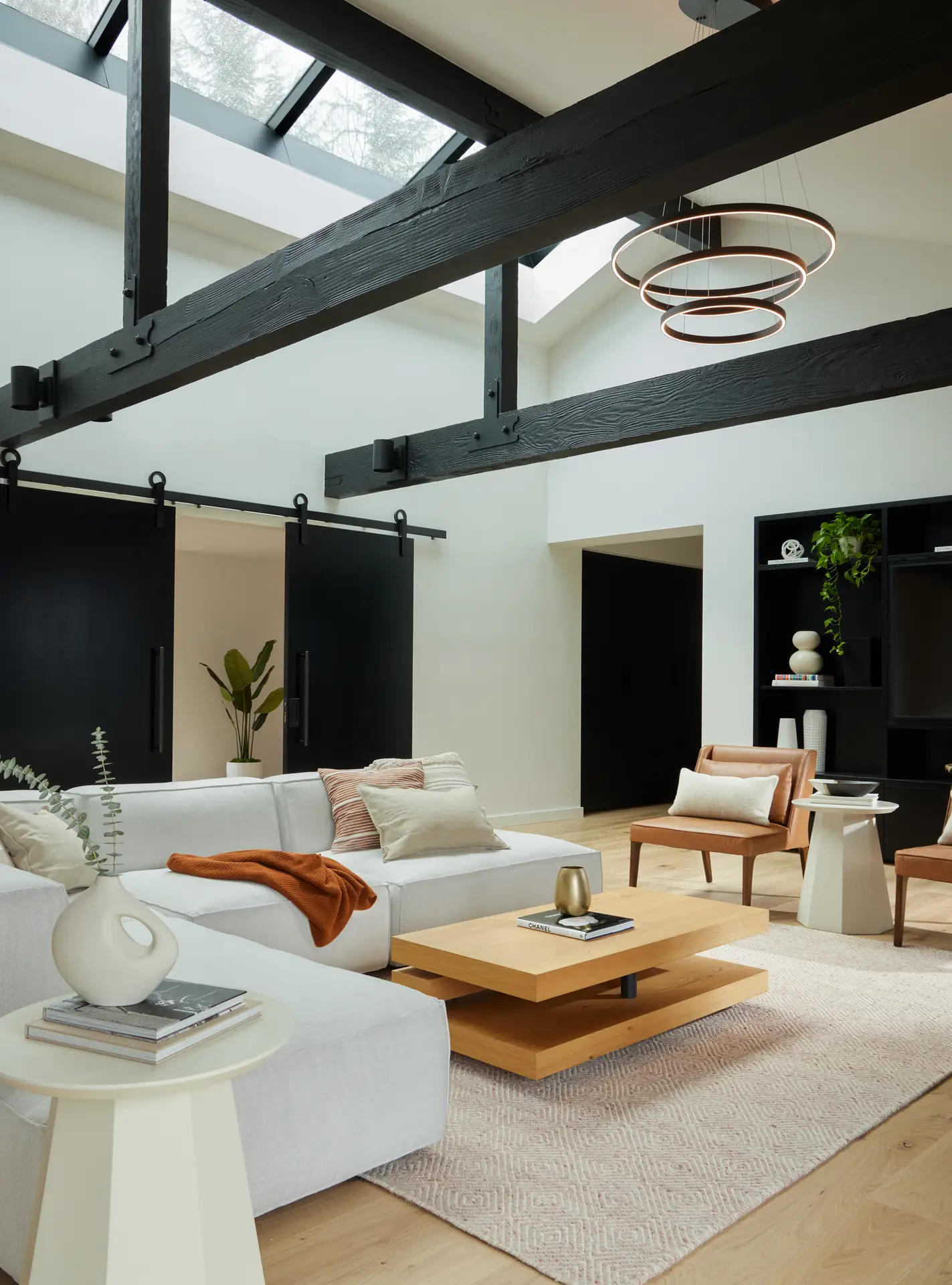 The vaulted ceiling exposed heavy-timber beams. painted matte black, providing a dramatic structural element. On the left two large black barn-style sliding doors on tracks offer a sleek, industrial way to transition between rooms. At the back of the room is a custom black floor-to-ceiling shelving unit provides storage and display space for ceramics and greenery, contrasting sharply with the crisp white walls.