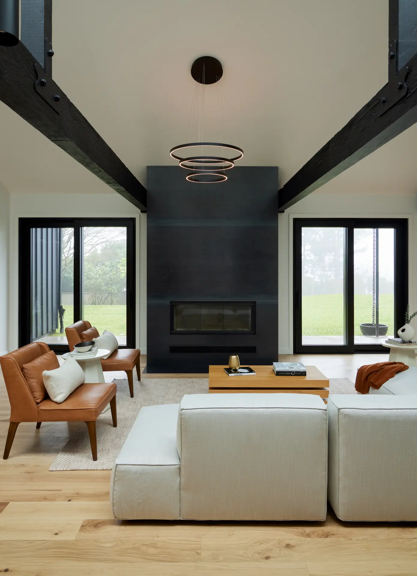 A front view of the custom metal clad fireplace. Two mid-century modern accent chairs, feature warm tan leather upholstery and tapered wooden legs. A low-profile, boxy modular sofa in a light oatmeal or beige fabric provides expansive seating. A simple, rectangular wooden coffee table with a natural finish sits in the center of the seating area.