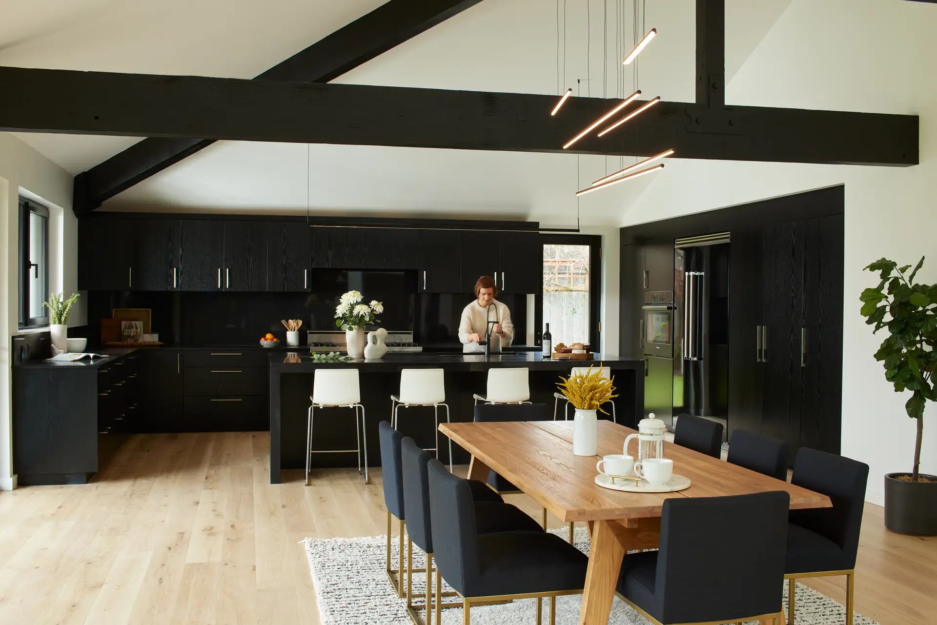 A modern kitchen and dining area of the Bridlemile Residence. Wide-plank white oak flooring adds organic warmth and balances the dark architectural elements. A large walnut dining table is paired with eight black upholstered chairs on a textured area rug. Four white Eames-style molded stools with chrome legs provide a crisp contrast against the black island in the background. A minimalist black linear LED pendant is suspended over the island, while a more sculptural multi-baton LED fixture hangs over the dining area.