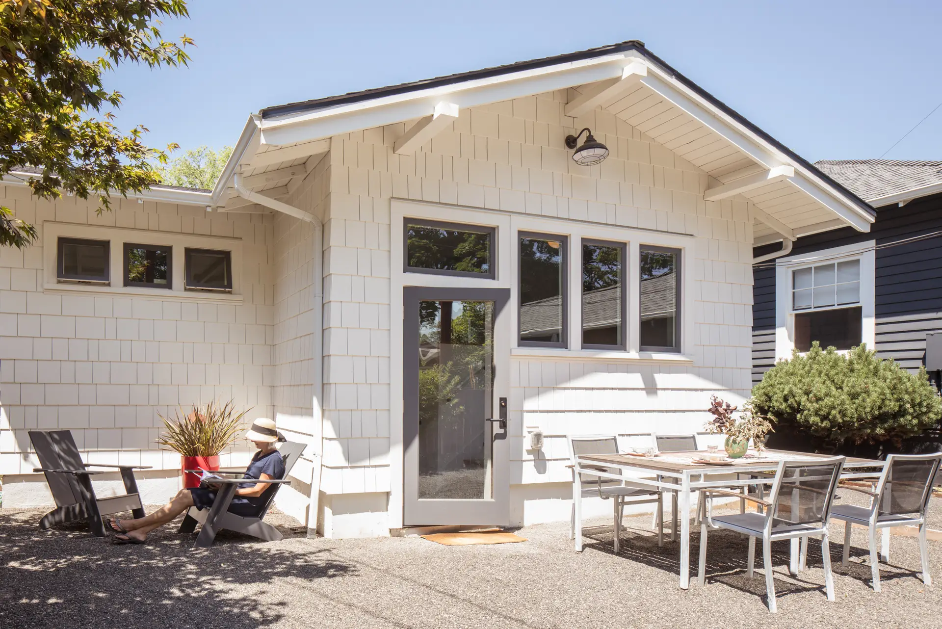 Little Larch exterior view, featuring a charming craftsman style home with white siding and thoughtfully placed windows. The foreground is comprised of a patio, patio table and chairs, and a person sitting in the left side of the frame.
