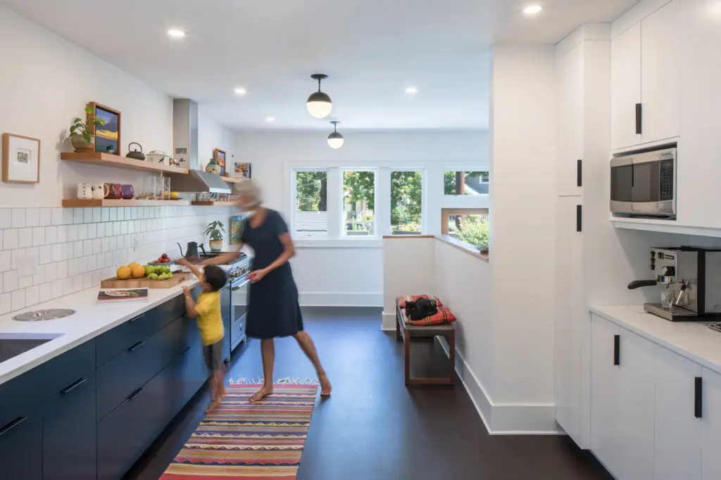 Little Larch kitchen, highlighting a beautiful galley style kitchen with dark brown floors and navy blue lower cabinets balanced out by light and bright tile and walls. Two Schoolhouse ceiling mount light fixtures create a focal point leading toward the windows beyond.