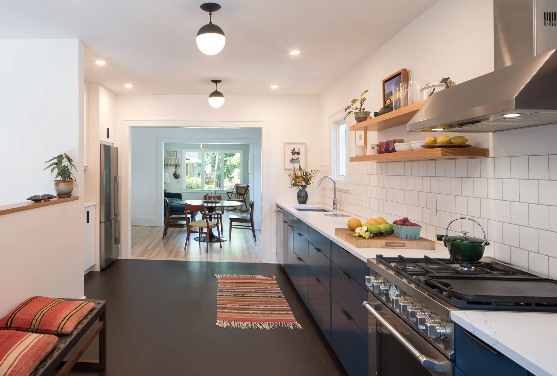 Little Larch kitchen, highlighting a beautiful galley style kitchen with dark brown floors and navy blue lower cabinets balanced out by light and bright tile and walls. Two Schoolhouse ceiling mount light fixtures create a focal point leading toward the dining room and windows beyond.